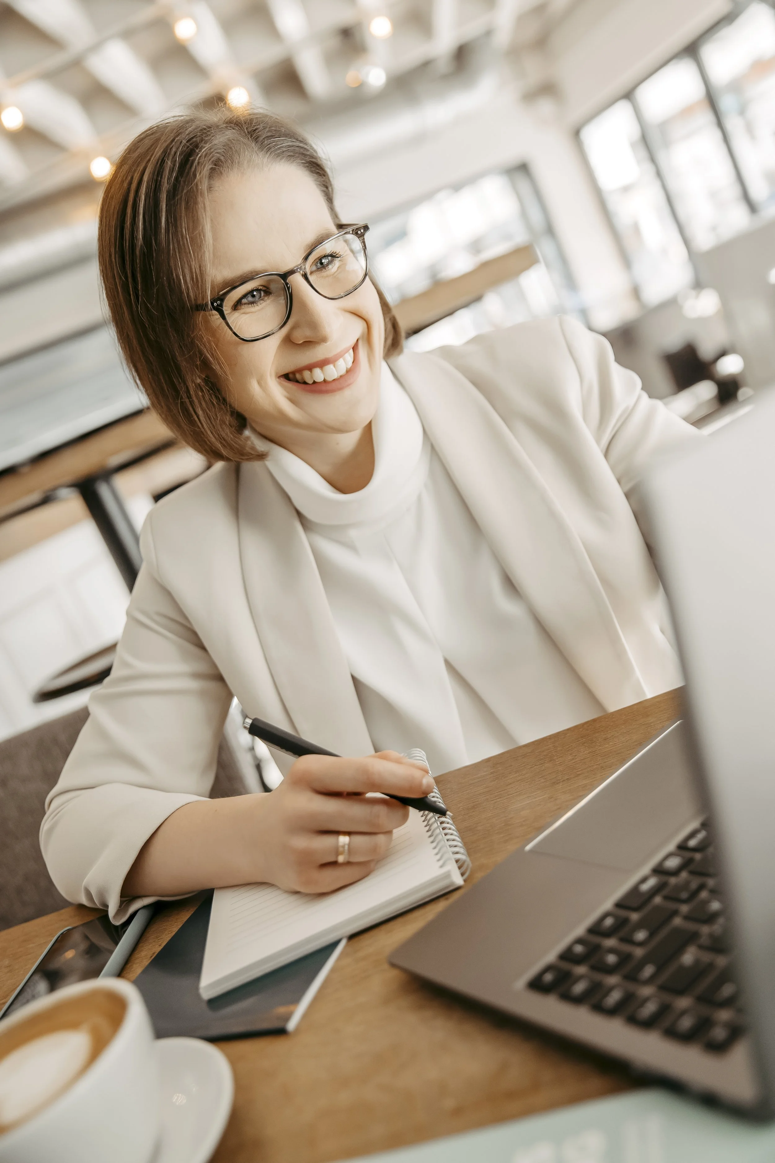 Eine lächelnde Frau mit Brille arbeitet in einem Café, schreibt in ein Notizbuch und hat einen Laptop vor sich.