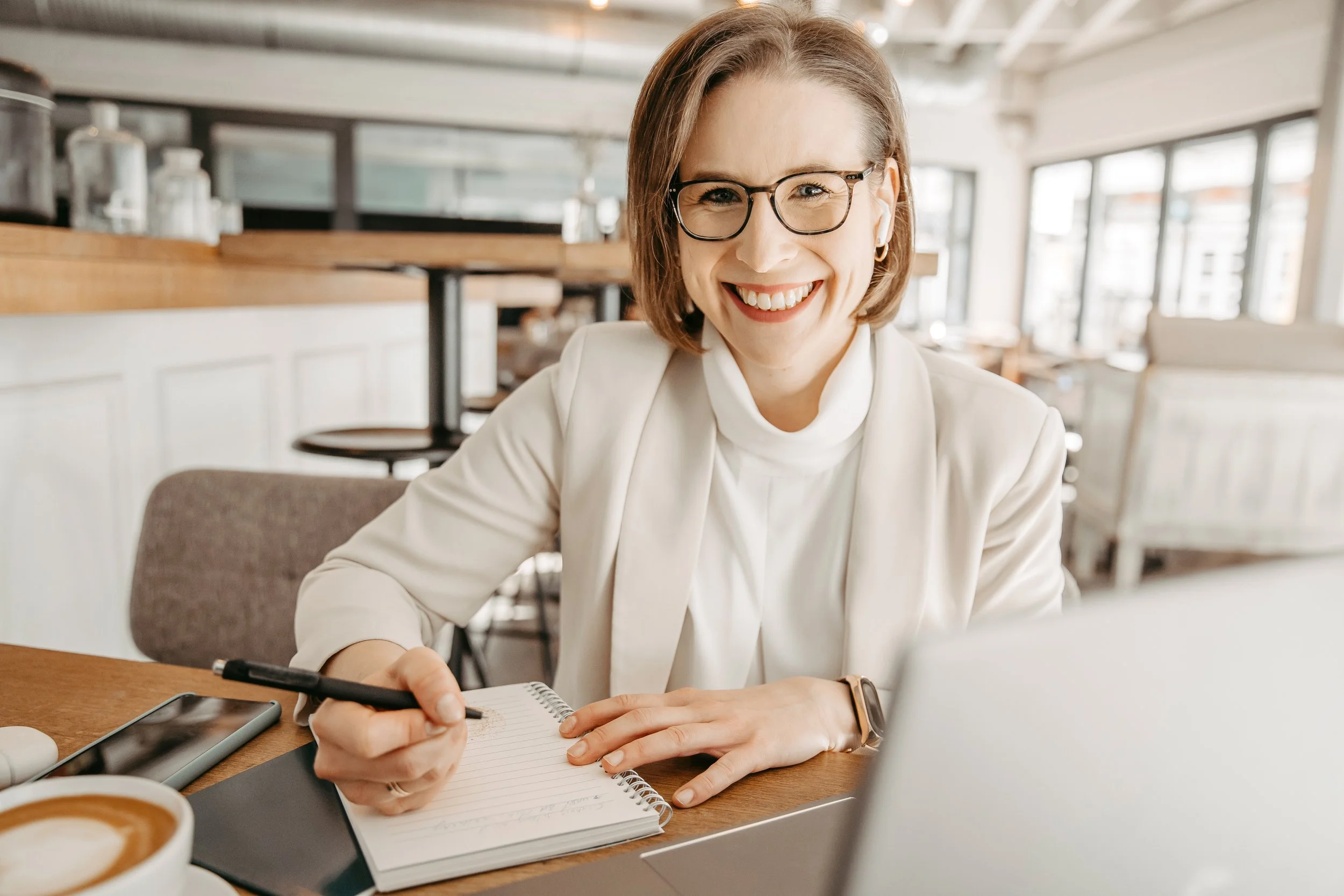 Lächelnde Frau mit Brille sitzt an einem Tisch im Café und schreibt in ein Notizbuch, während sie einen Laptop benutzt.