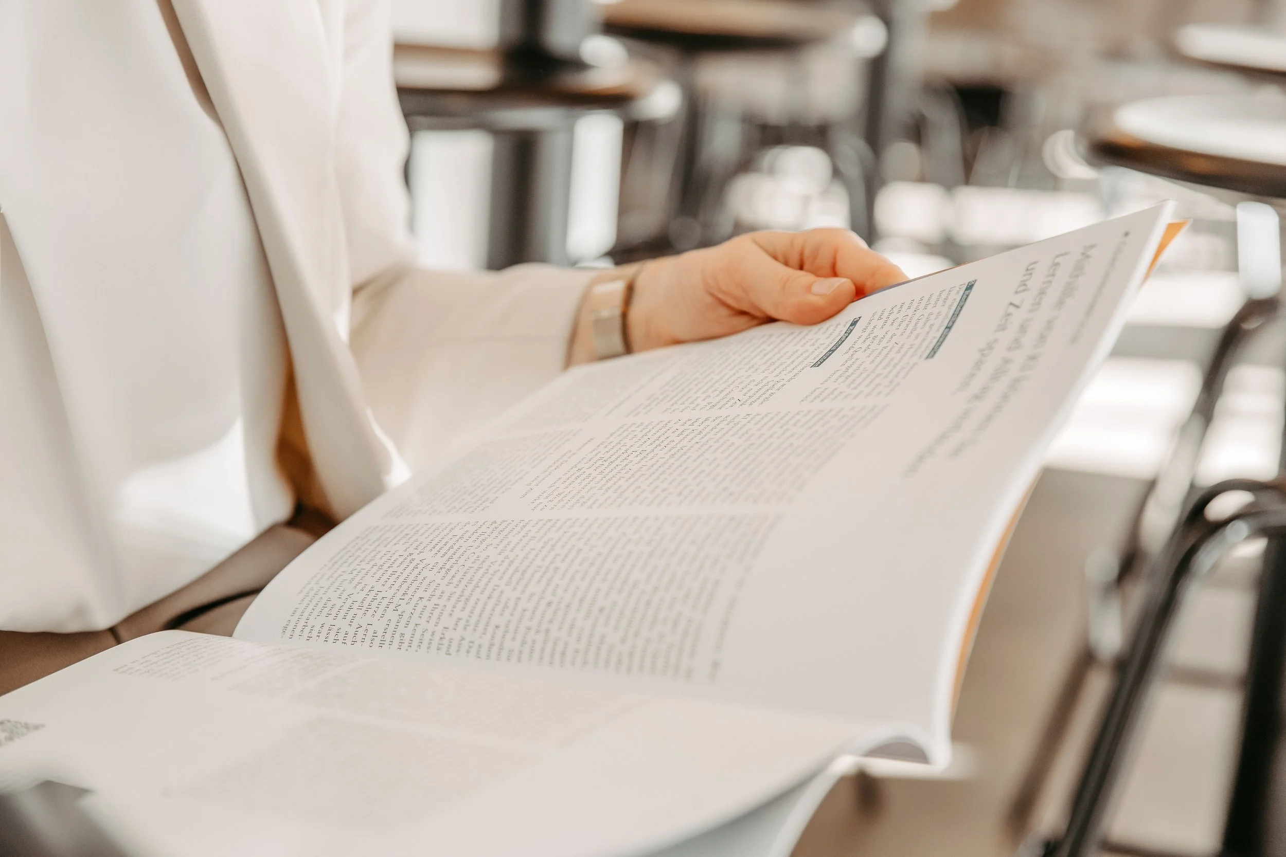 Eine Person liest eine Zeitung oder ein Magazin in einem öffentlichen Raum, vermutlich in einem Café oder einer Bibliothek.