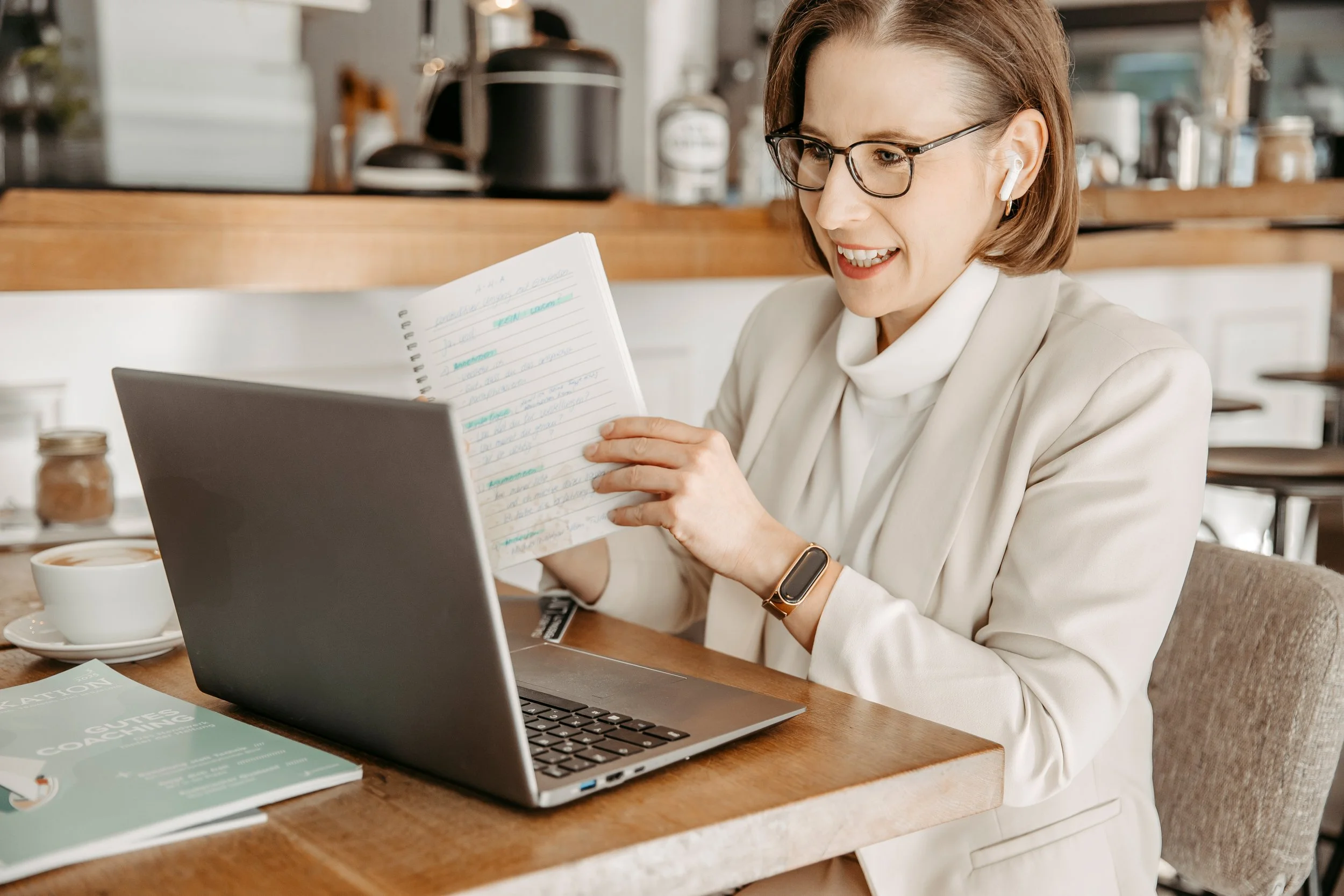 Eine Frau sitzt an einem Holztablettisch in einem Café, trägt eine Brille und Ohrhörer, liest eine Notizseite und hat einen Laptop, eine Kaffeetasse und eine Zeitschrift vor sich.