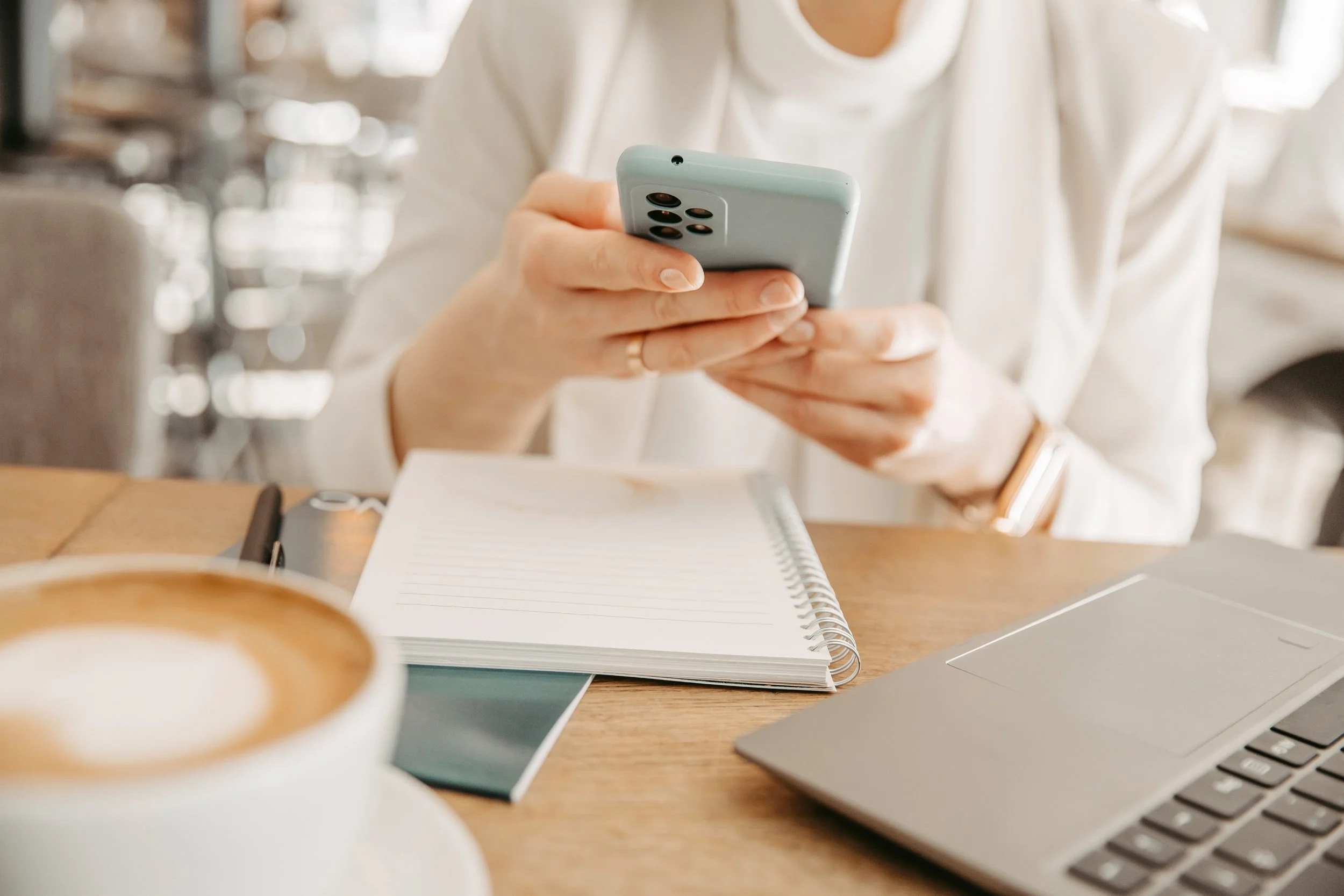 Person verwendet Smartphone an einem Tisch mit Notizbuch, Laptop, Stift und Kaffee in einer Cafeteria.