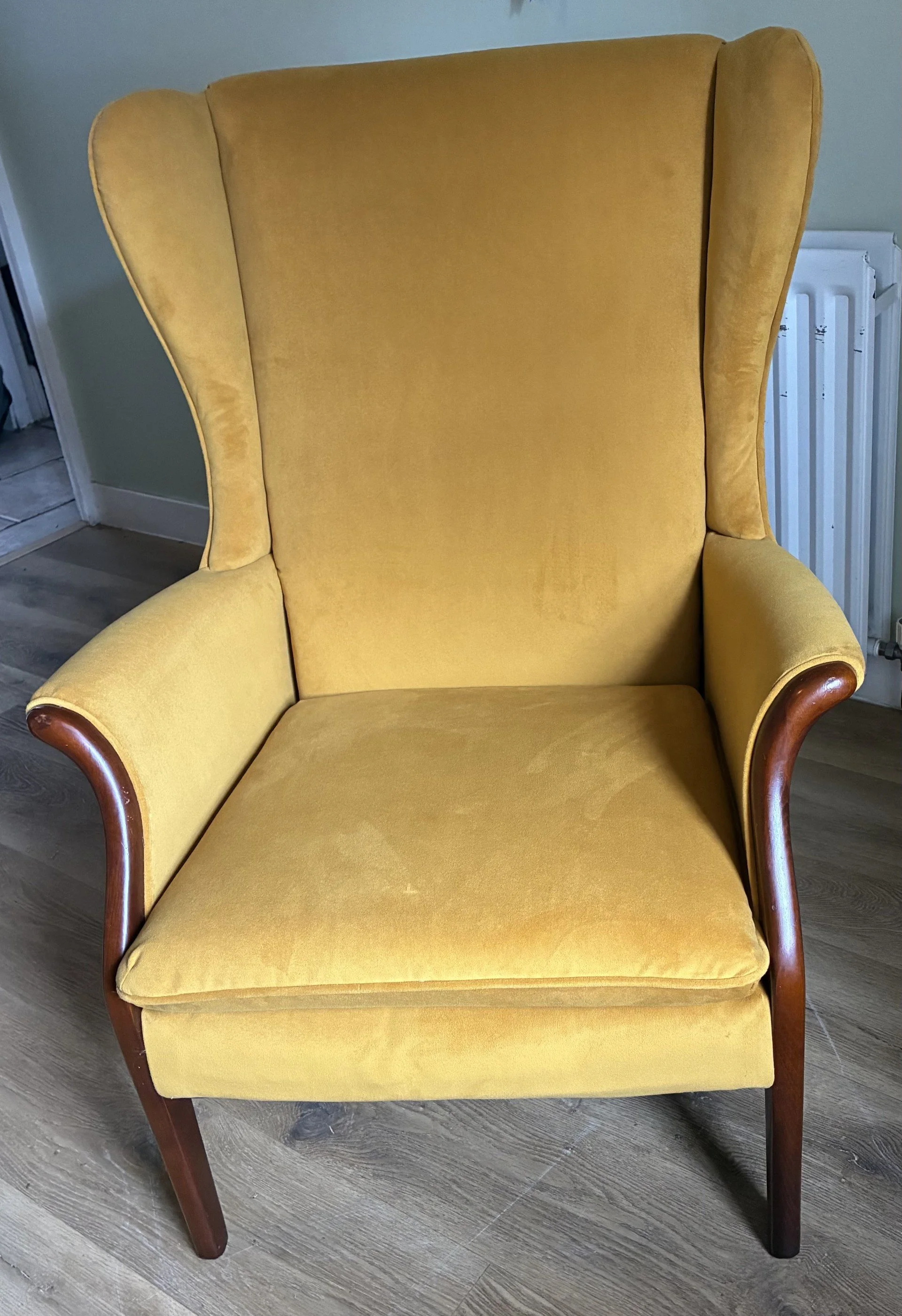 A yellow upholstered armchair with wooden arms, situated on a hardwood floor next to a radiator.