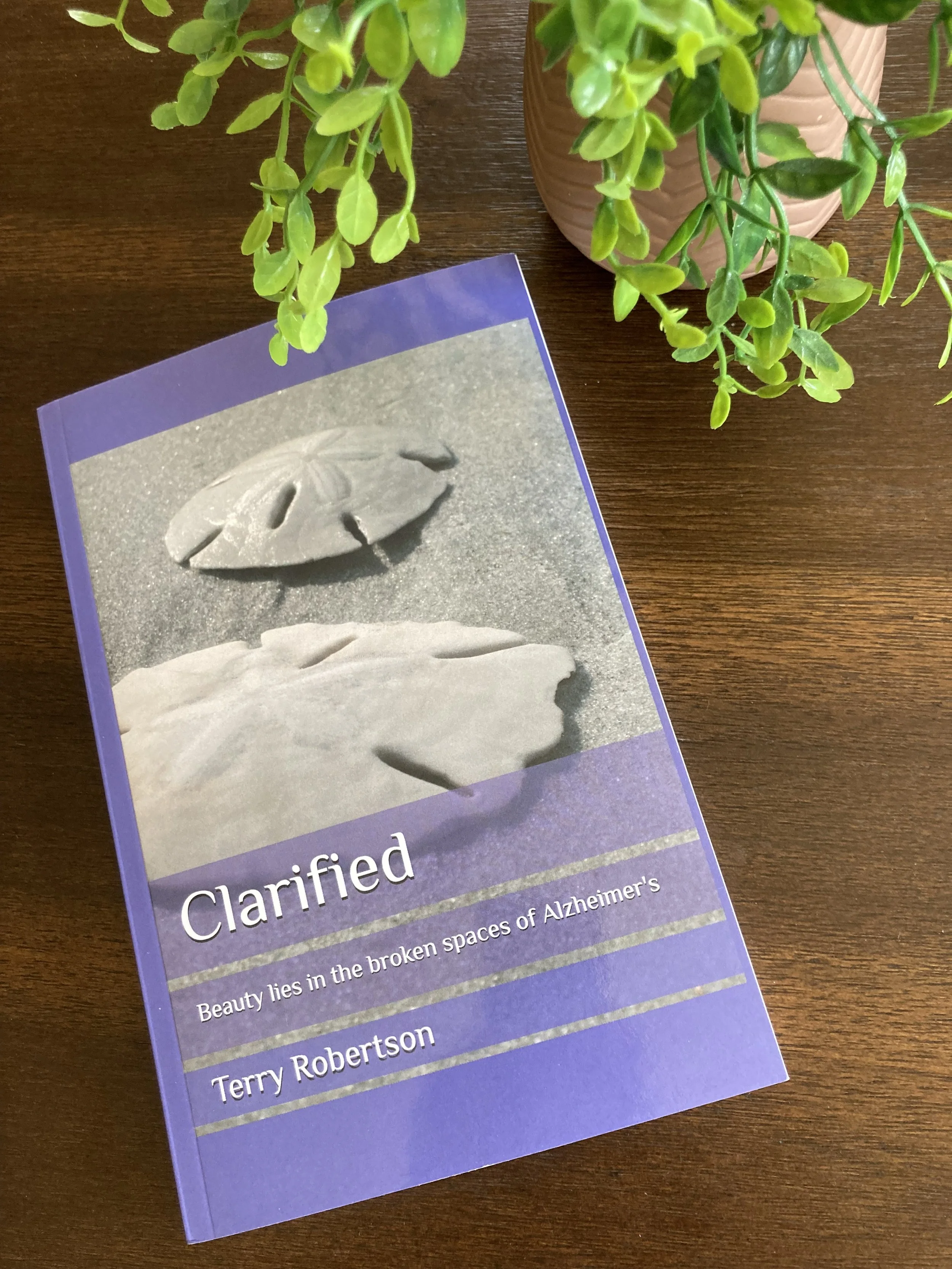 A book titled 'Clarified' by Terry Robertson resting on a wooden surface near a potted plant with green leaves.