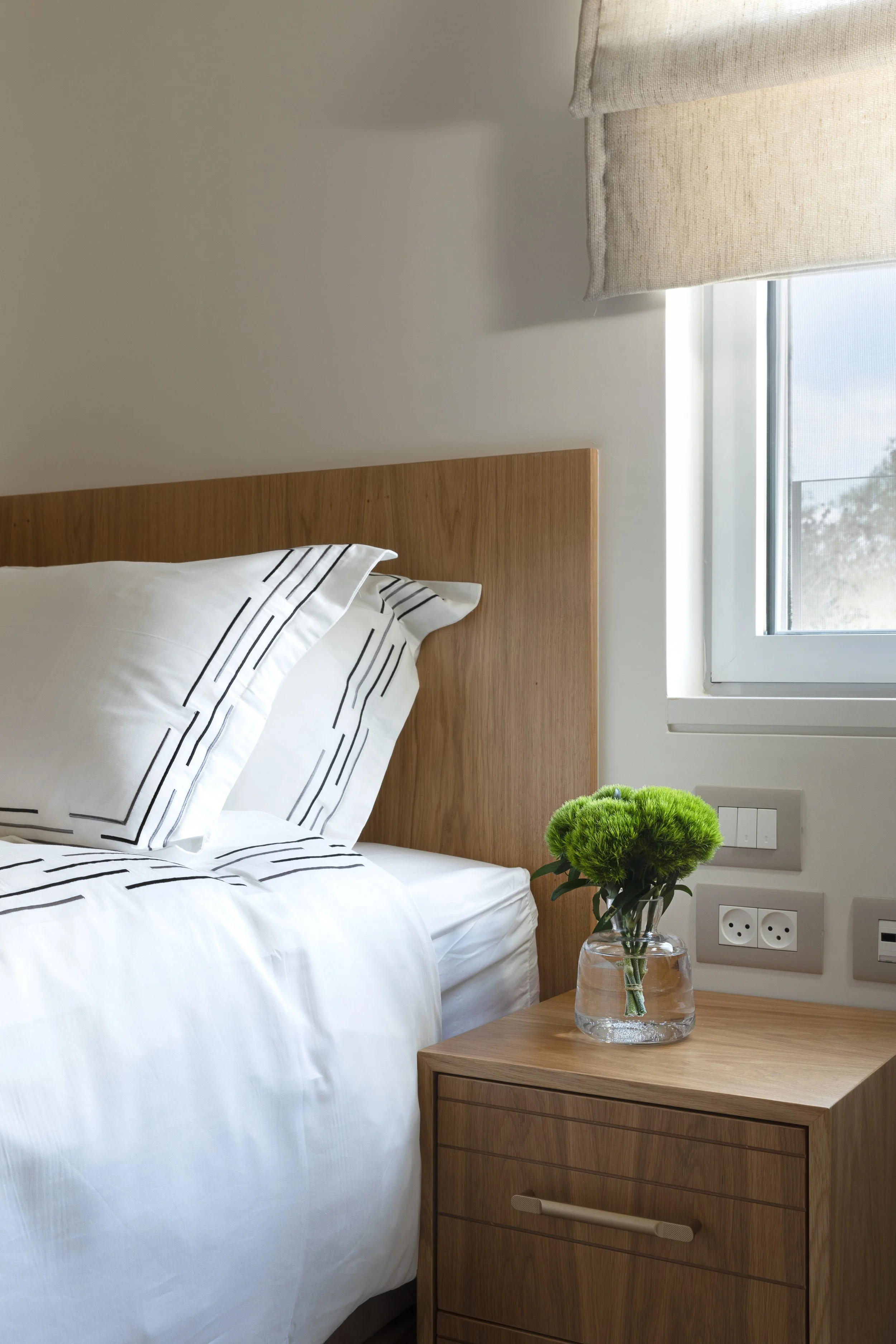 A bedroom with a wooden headboard, white bedding with black striped accents, a nightstand with a glass vase of green flowers, a beige window shade, and electrical outlets on the wall.
