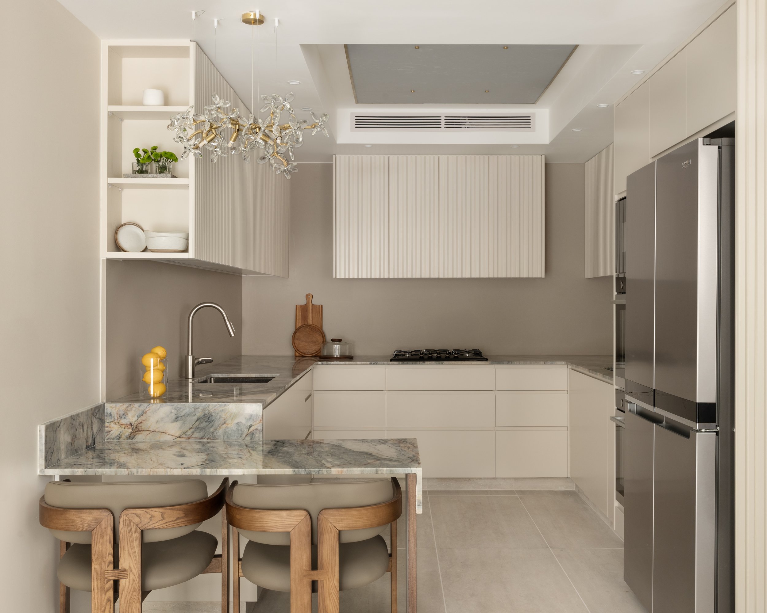 Modern kitchen with a marble counter, tan chairs, a white backsplash, and upper white cabinets. A decorative chandelier hangs from the ceiling, and there are small plants and bowls on the shelves.