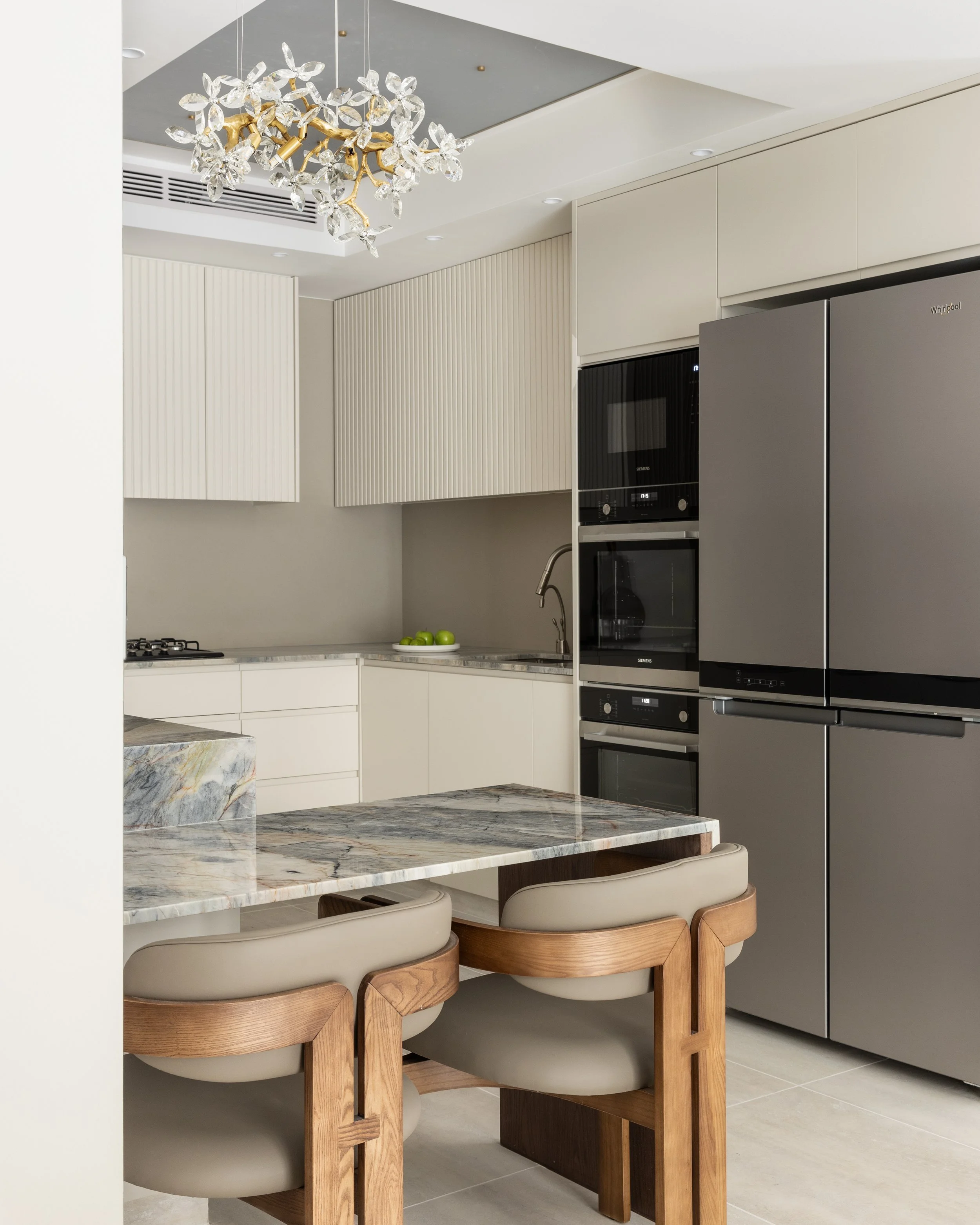 Modern kitchen with a marble-topped island, wooden chairs, white cabinetry, a stainless steel refrigerator, and a chandelier with leaf-shaped crystals hanging from a gold branch.