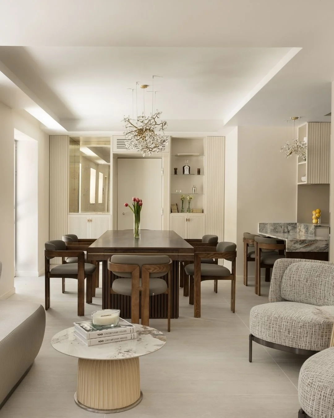 Modern dining and living room with white walls, beige flooring, a round marble coffee table with books and a candle, a large wooden dining table with six chairs, a vase with pink tulips, built-in shelves with decor, and a curved armchair.