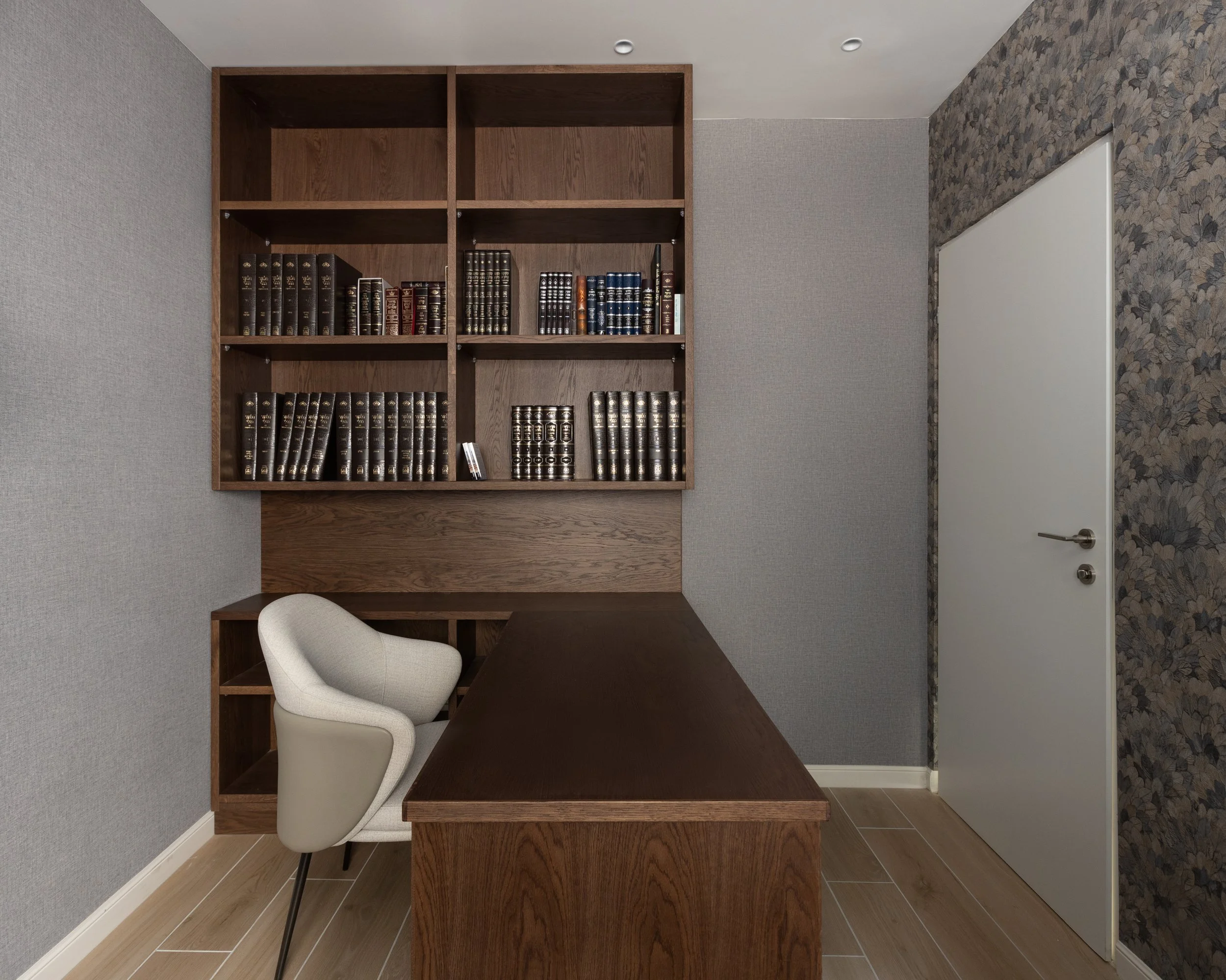 An office with a wooden desk and a white chair, a wooden bookshelf with leather-bound books, gray textured wall, and a door with a silver handle.