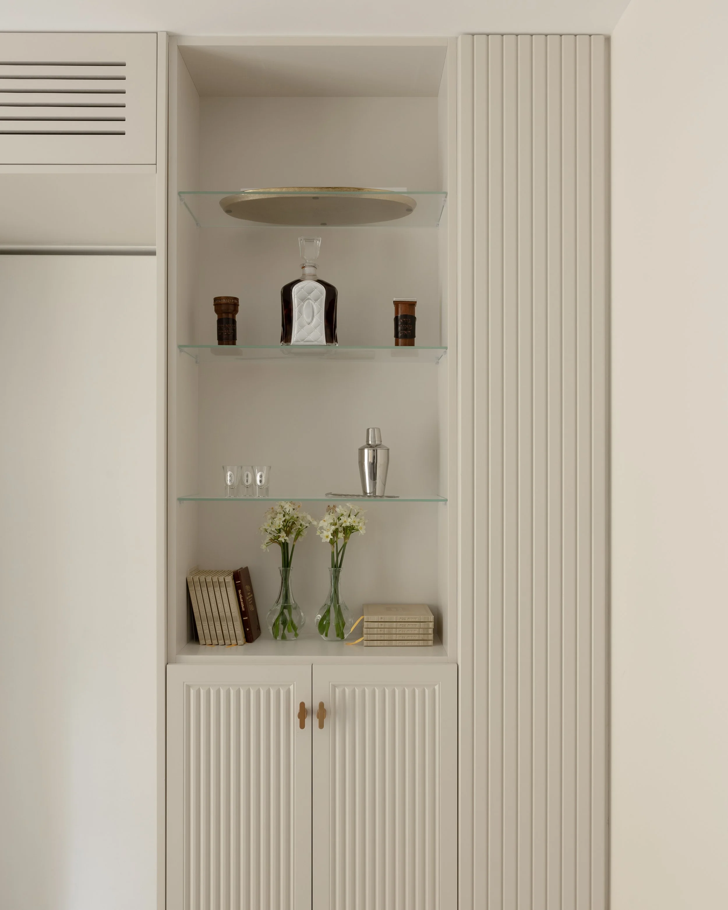 White built-in cabinet with four glass shelves displaying decorative items including books, flowers, and glassware.