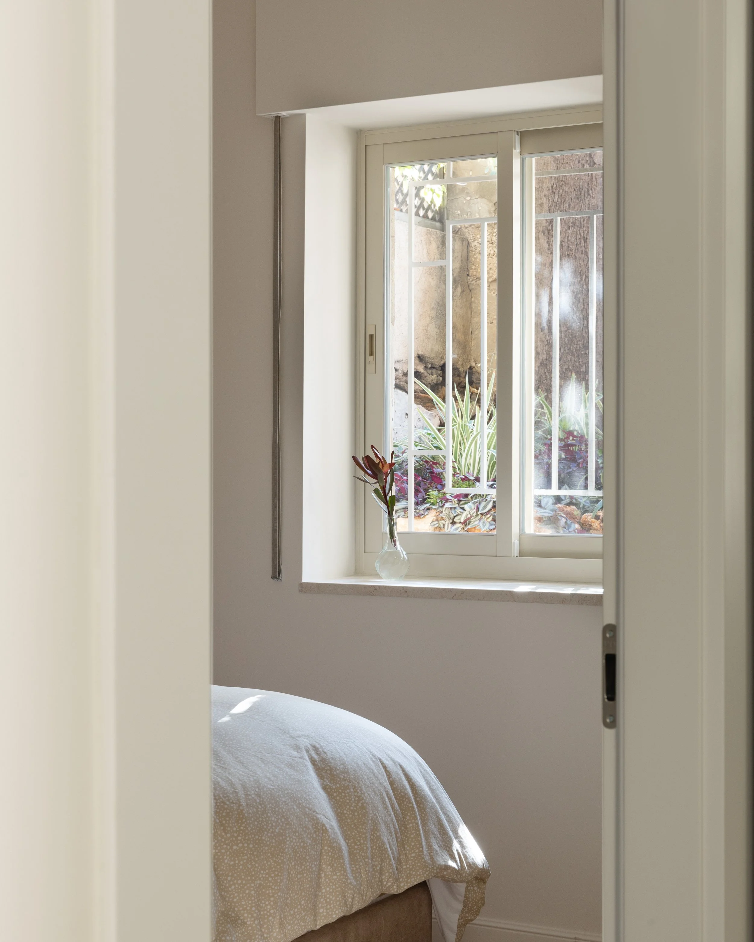 View of a bedroom with a bed covered in a light-colored duvet near a window, with a clear vase containing flowers on the windowsill, interior has neutral tones and natural light.