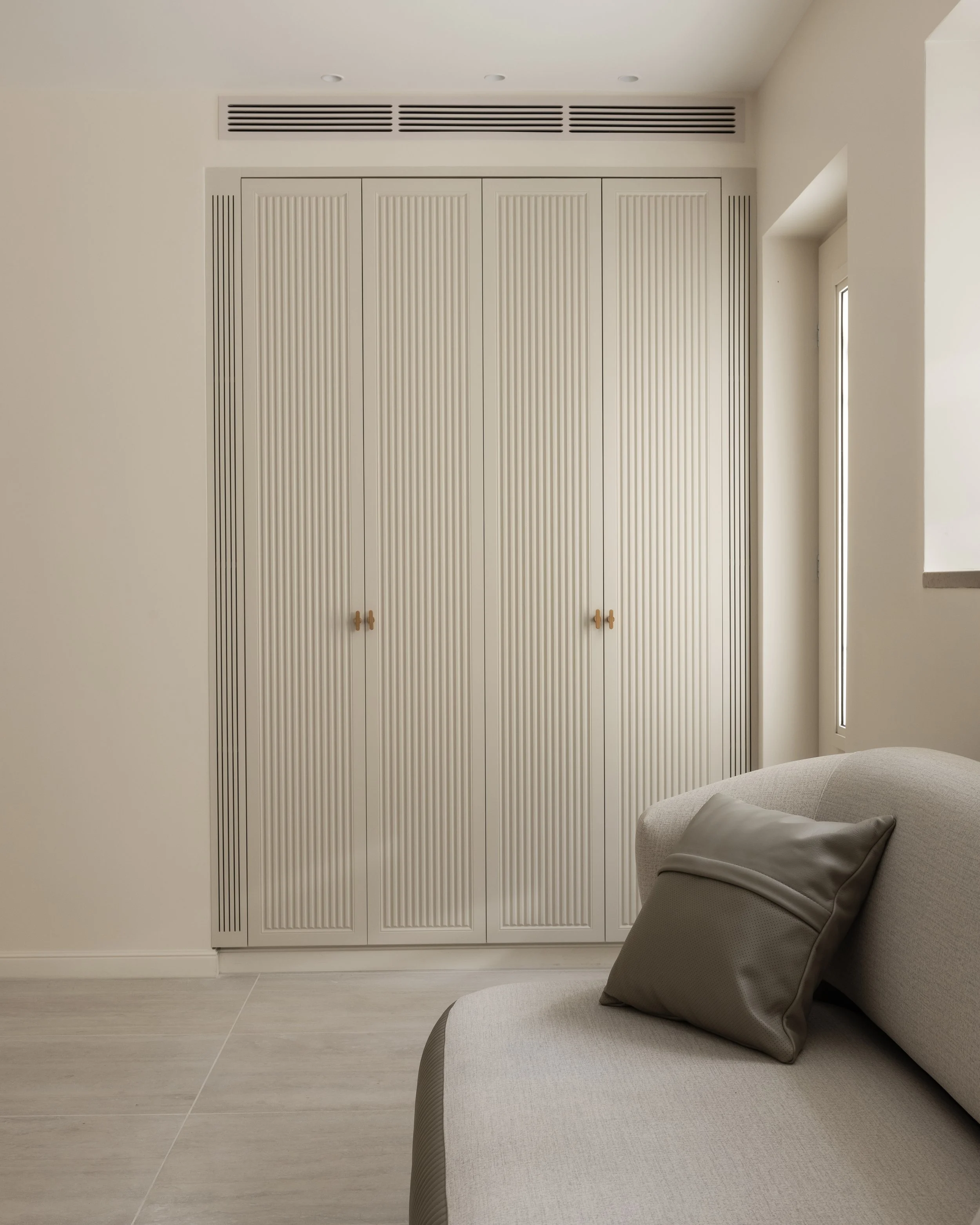 A beige lounge chair with a pillow in the corner of a room, near a white closet with louvered doors and an air vent above. Part of a window is visible on the right.
