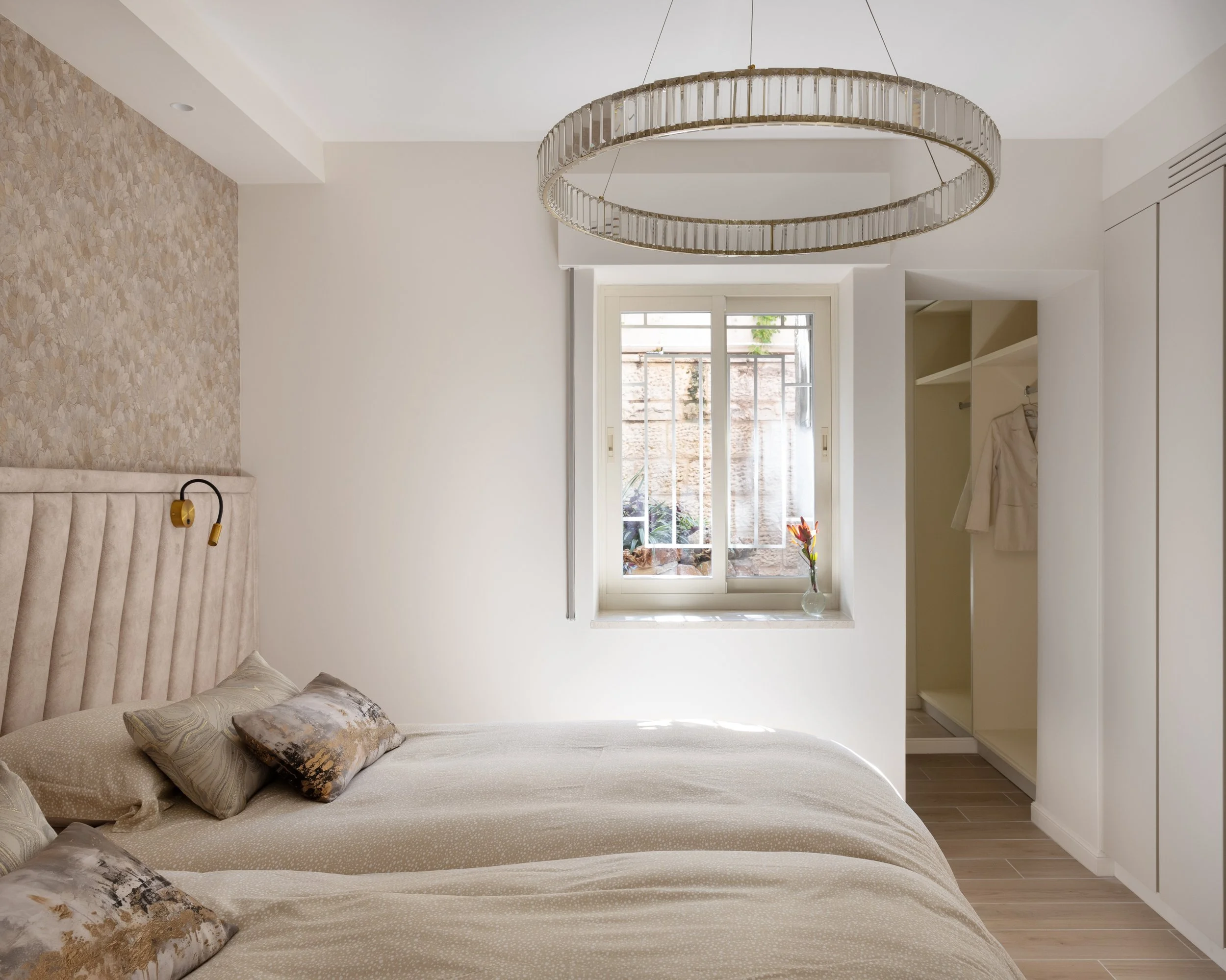 A bedroom with a large window, beige bed with multiple pillows, a patterned wallpaper on the headboard wall, a modern chandelier, and a closet.