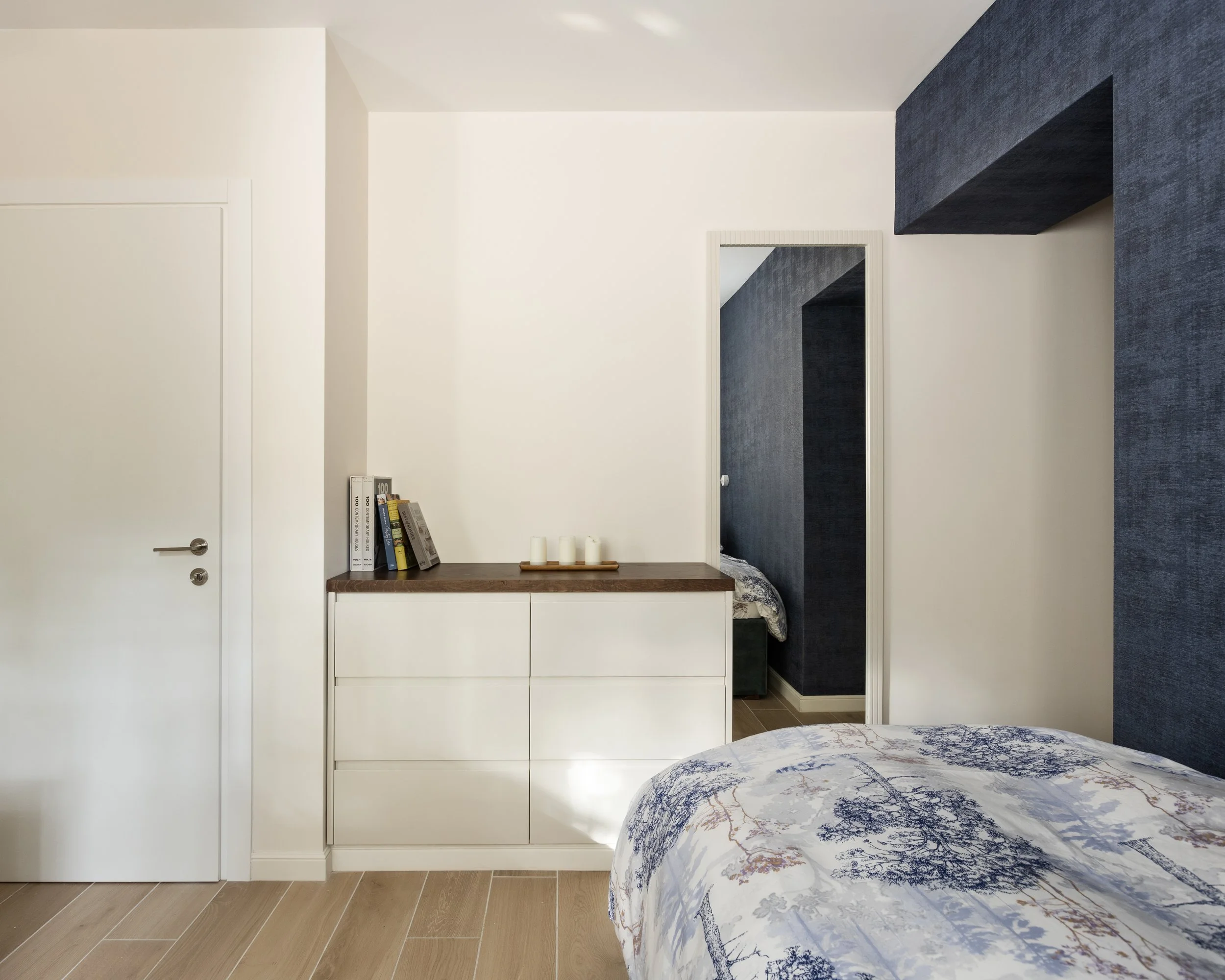 Interior view of a modern bedroom with white walls, dark blue textured accent wall, and a wooden dresser with books and candles. There is a mirror on the wall reflecting part of the room, and a marble-patterned bed in the foreground.