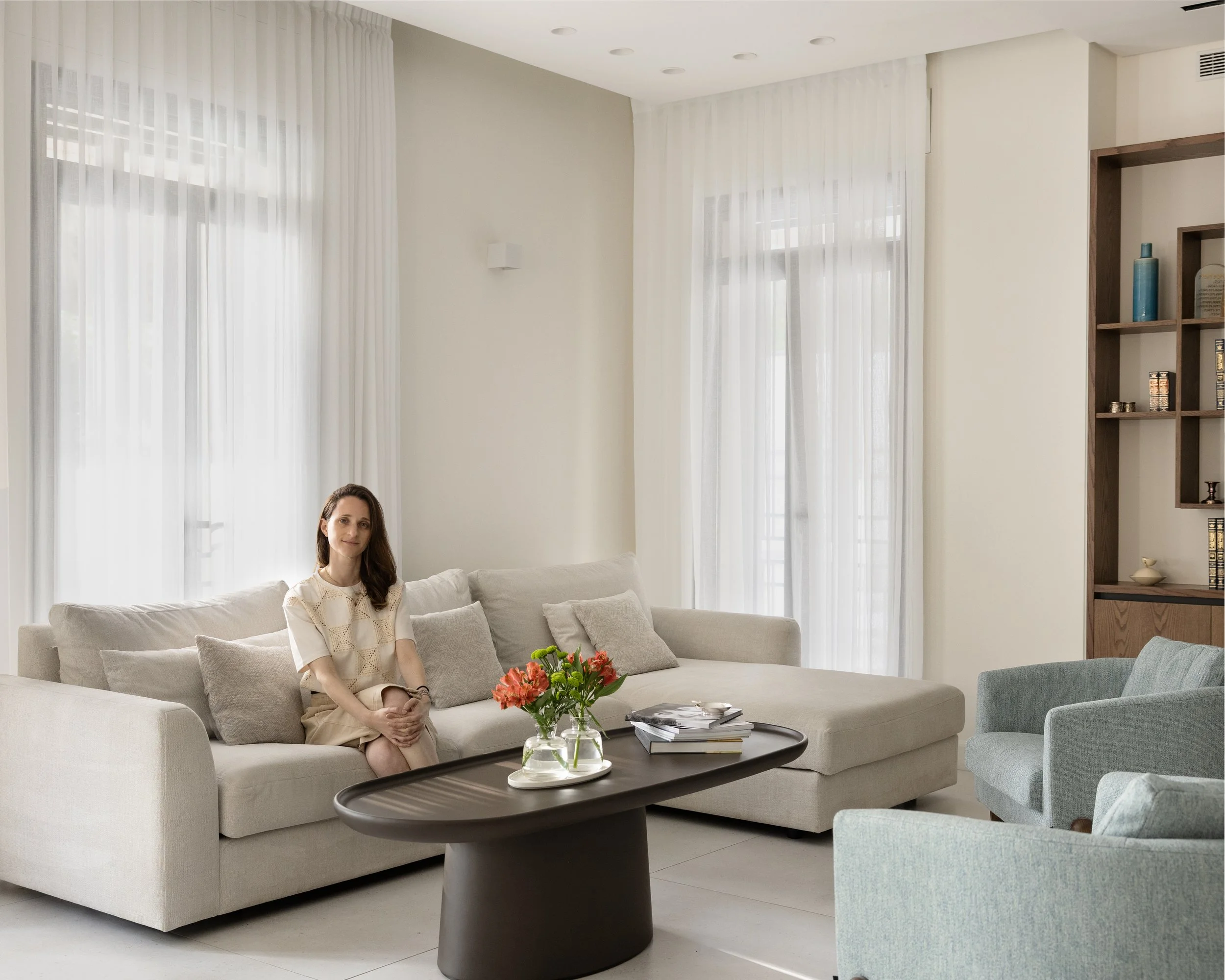 A woman sitting on a beige sofa in a living room with white curtains and a round coffee table with flowers and books.