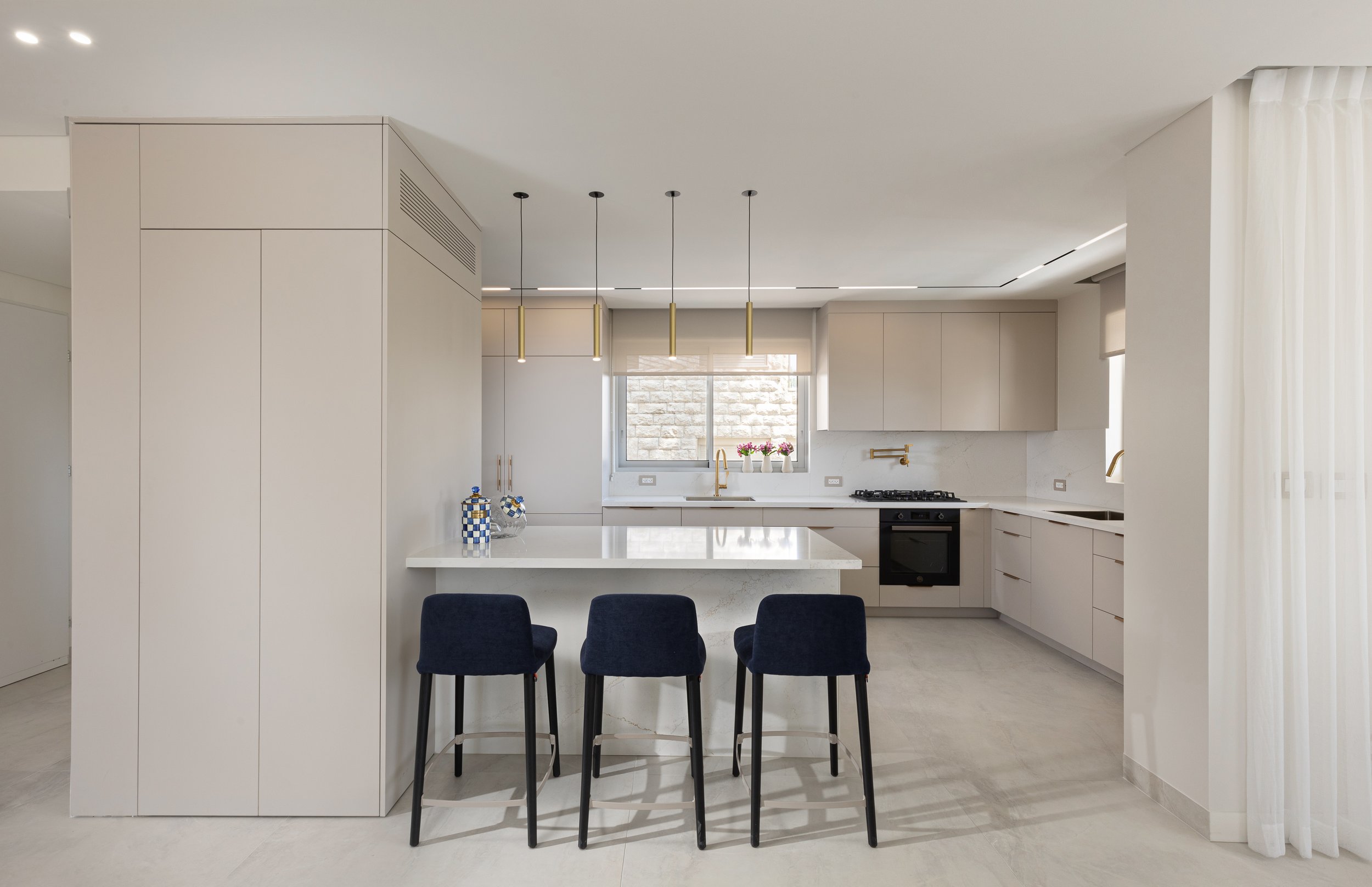 Modern kitchen with white cabinets, a white countertop island, black chairs, a window with natural light, and gold pendant lights.