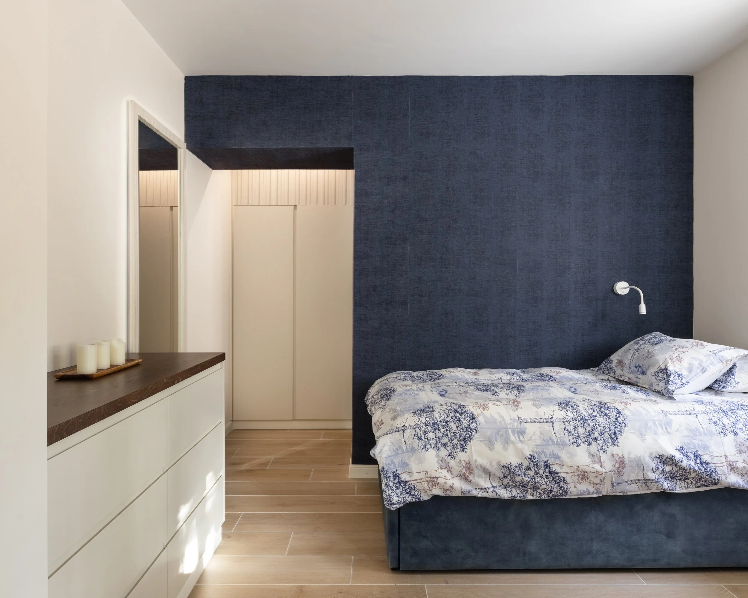 A bedroom with a blue textured headboard, a patterned bedspread, a white wall, and a dark blue accent wall. There is a built-in dresser with three white drawers and a tray with white candles.