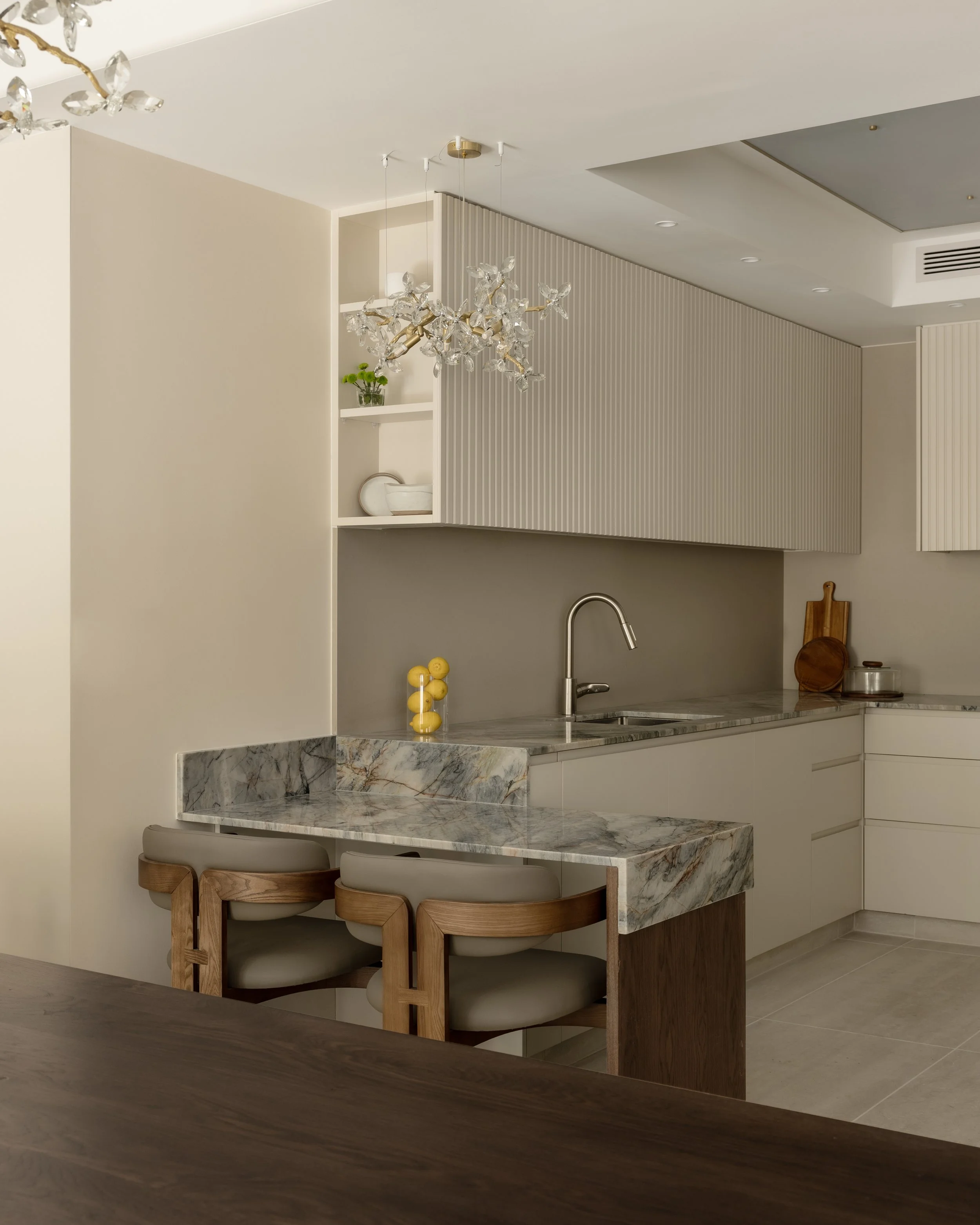 Modern kitchen with marble countertop, wooden stools, and a pendant chandelier with crystal accents.