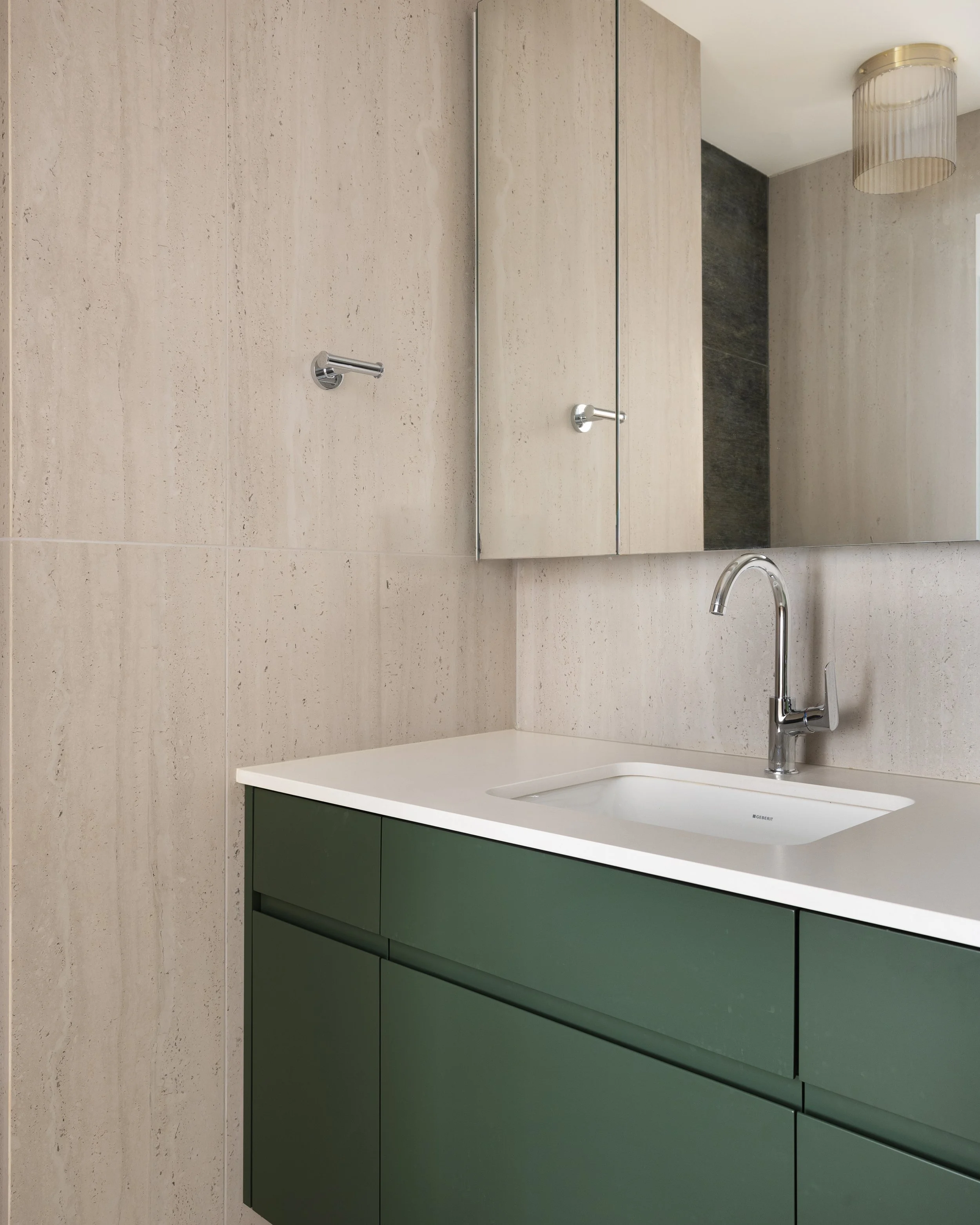 Modern bathroom with beige tiled wall, green vanity with white countertop, white sink, chrome faucet, large mirror, and a beige ceiling light fixture.