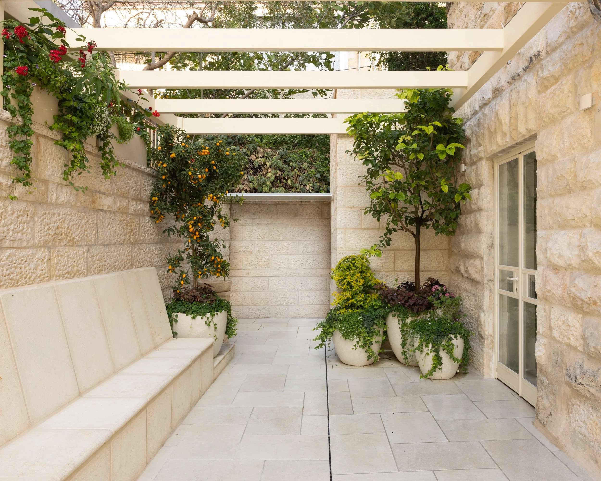 A small outdoor patio area with a built-in bench along the left wall, surrounded by potted plants and trees, with stone walls and a pergola above.
