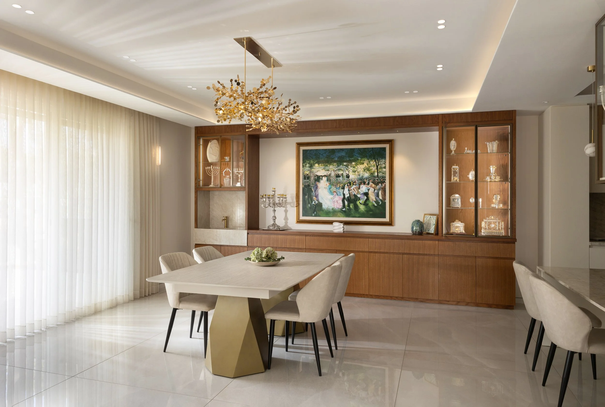 Modern dining room with white table, beige chairs, wooden built-in shelves, artwork, and a chandelier.