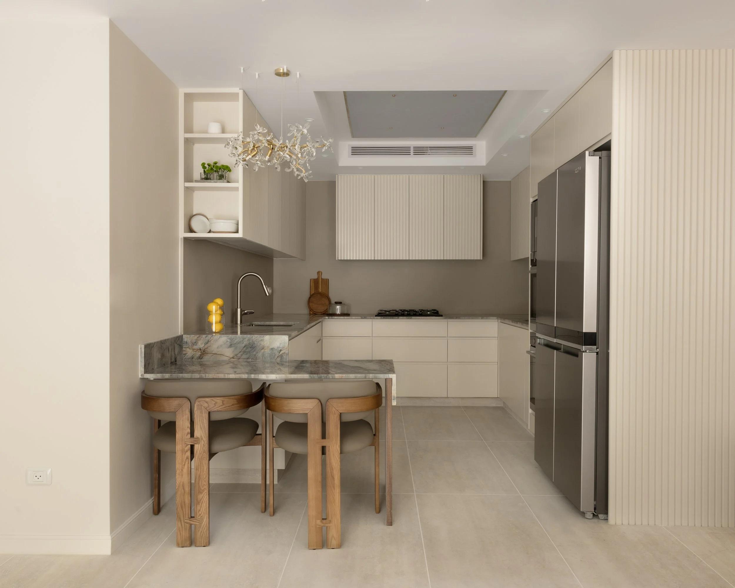Kitchen with cream cabinets, stainless steel refrigerator, marble island with stools, open shelving with dishes and plants, chandelier, beige floor tiles, and a blue ceiling detail.