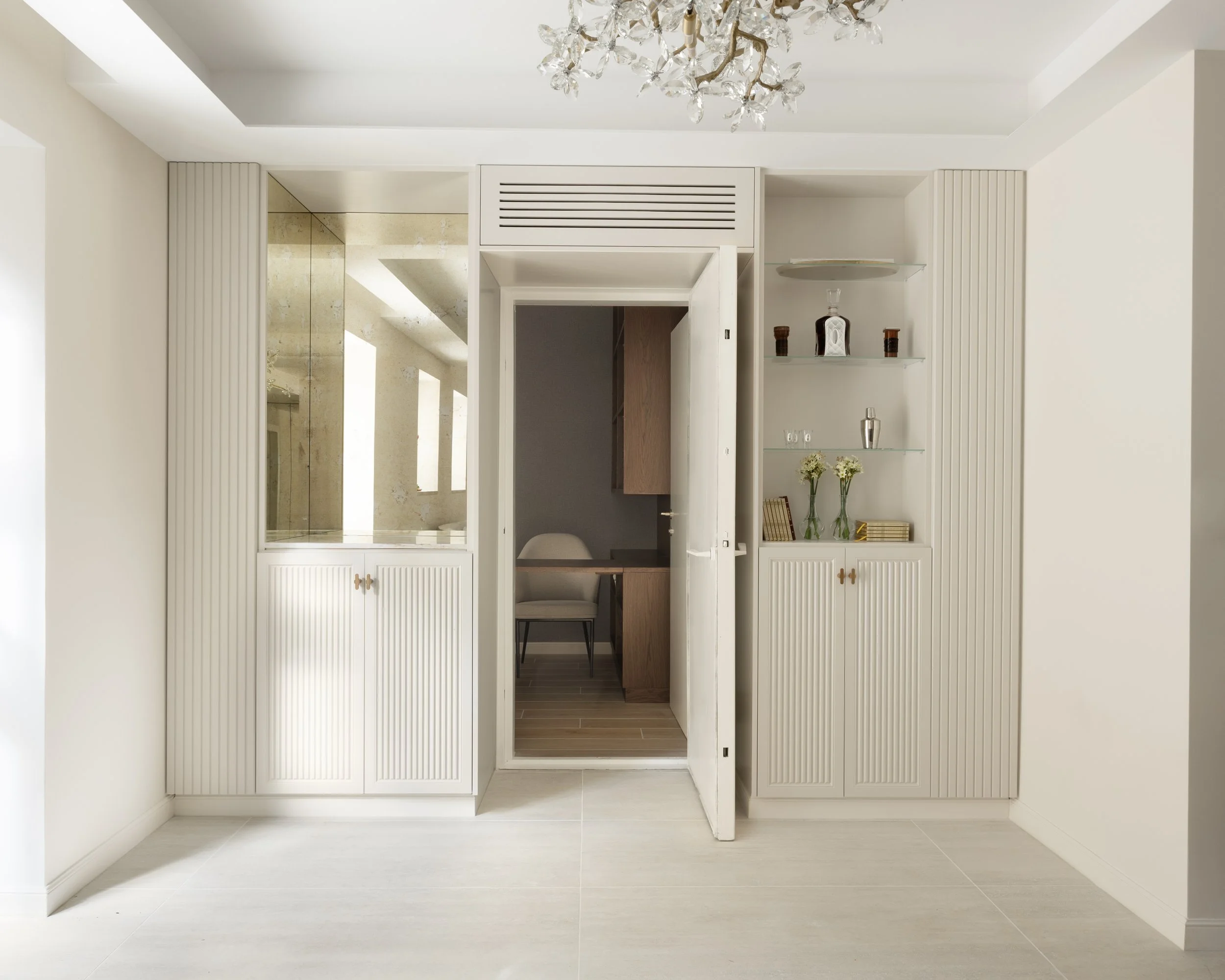 White built-in cabinet with glass shelves, decorative items, and flowers, with a doorway leading to a bathroom visible through a mirror reflection, white walls, and a ceiling chandelier.