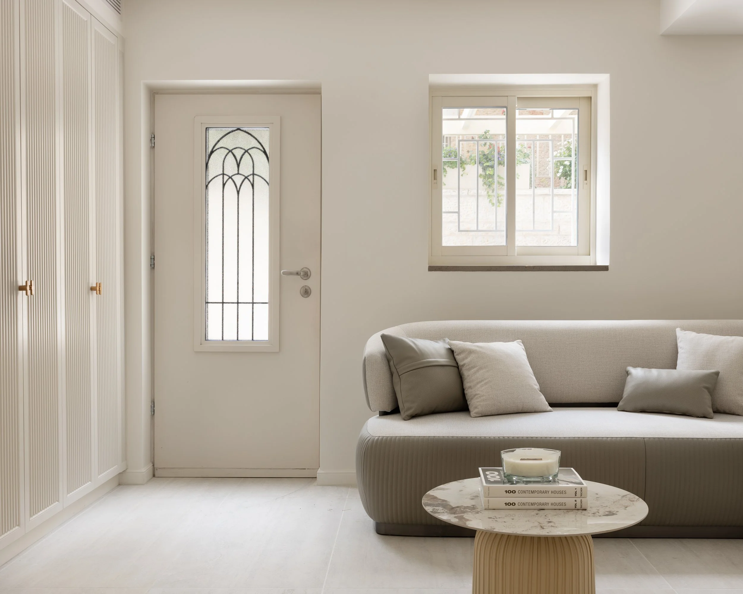 A minimalist living room with white walls, a large window with metal bars, a cream sofa with pillows, a white wardrobe with wooden handles, a round marble side table with books, and a candle.