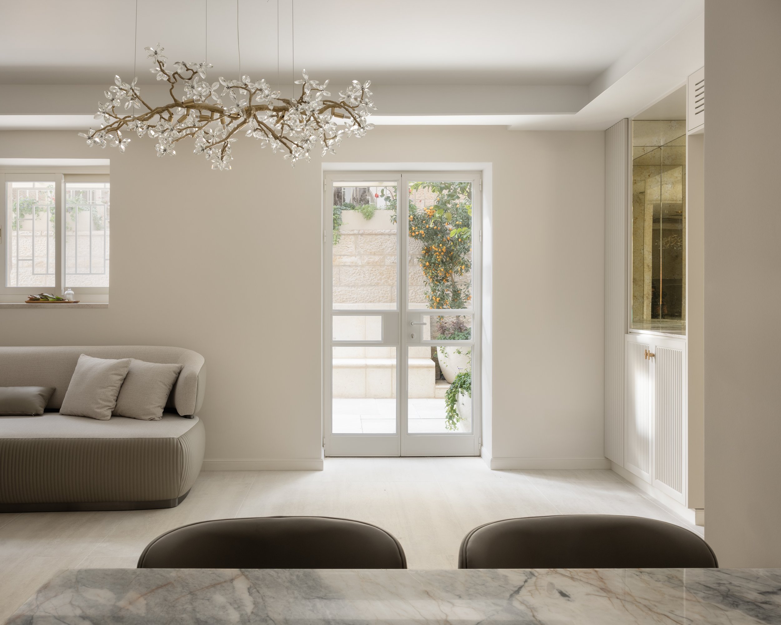 Living room with a white sofa, large window with a garden view, and a glass door leading outside. Modern chandelier with crystal accents hangs from the ceiling.