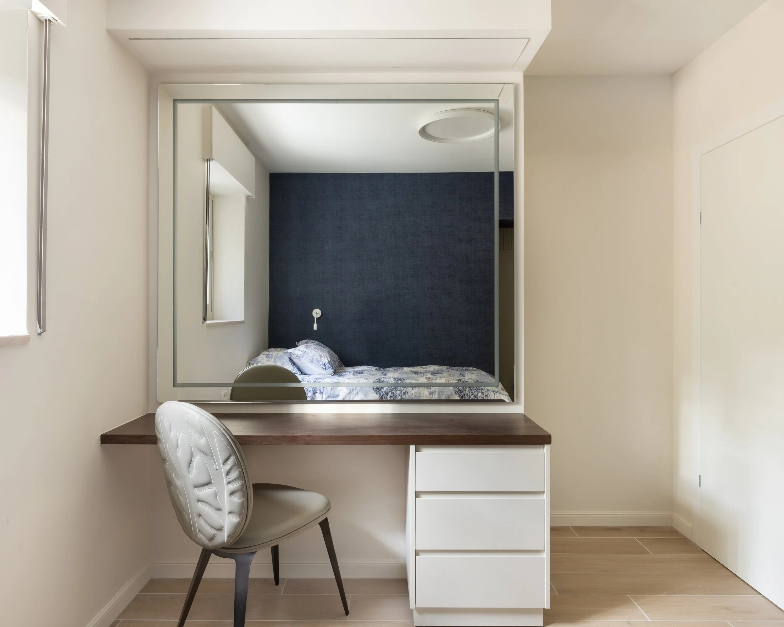 Small bedroom with white walls and wooden floors, a single bed with a floral blanket and pillows placed against a dark blue accent wall, a white desk underneath the bed, and a white chair with a patterned backrest in front of the desk, small window t