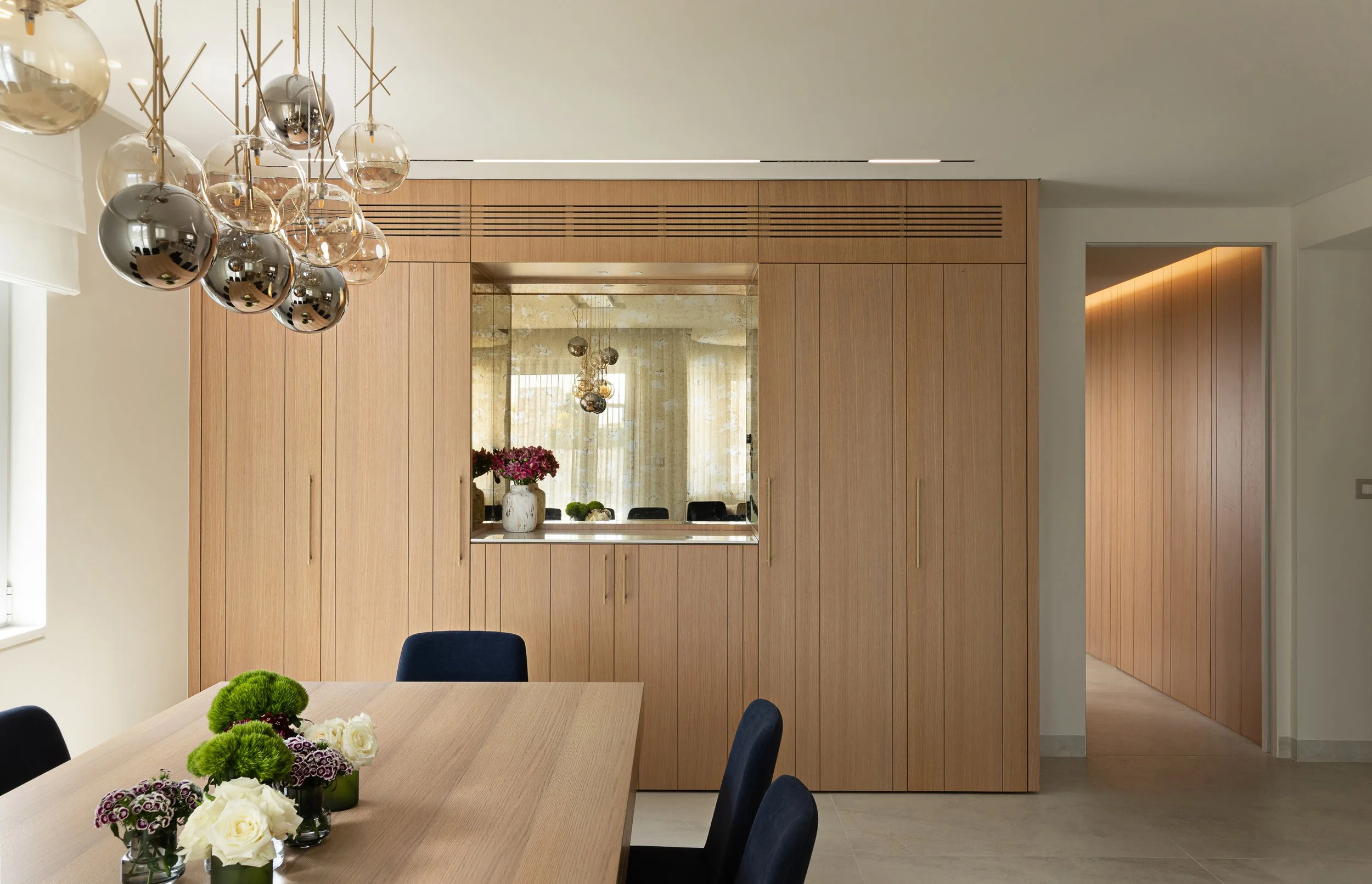Modern dining room with a light wood table, floral centerpieces, and black chairs. The background features wood-paneled walls, a mirror, and a built-in window. Hanging glass sphere light fixtures are above the table.