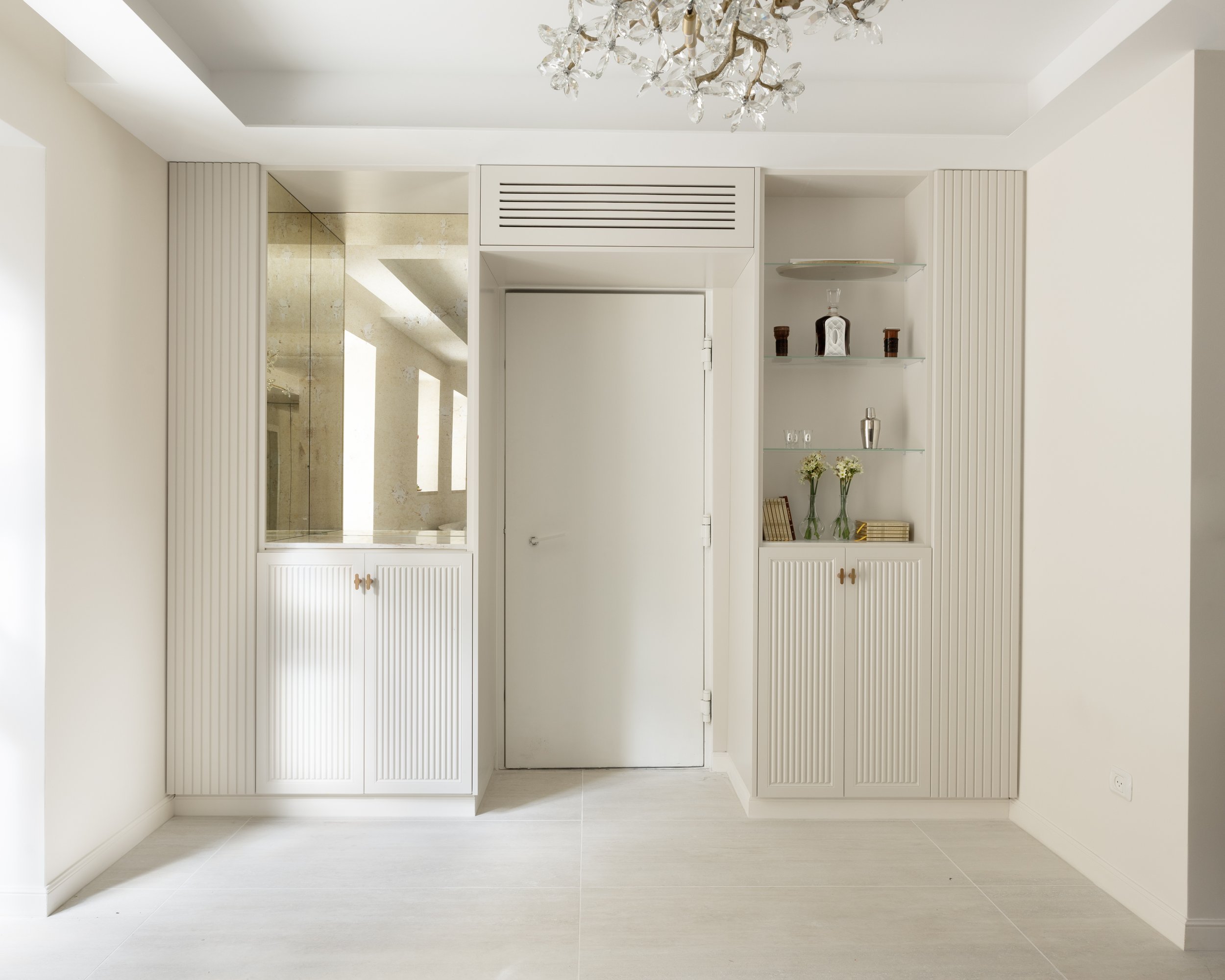 A white built-in cabinet with vertical grooves flanking a door, located in a minimal, bright room with white walls and tiled flooring. A glass-paneled door opening to a bathroom or shower area is visible on the left.