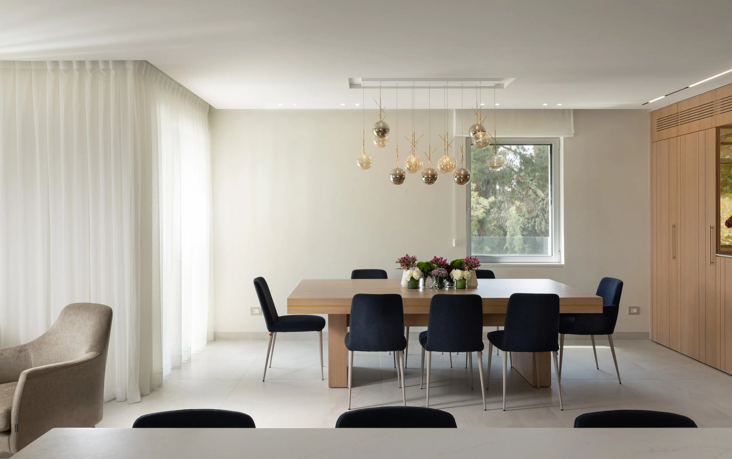 Modern dining room with a wooden table, six black chairs, a floral centerpiece, a large window, white curtains, and a modern chandelier with hanging glass globes.