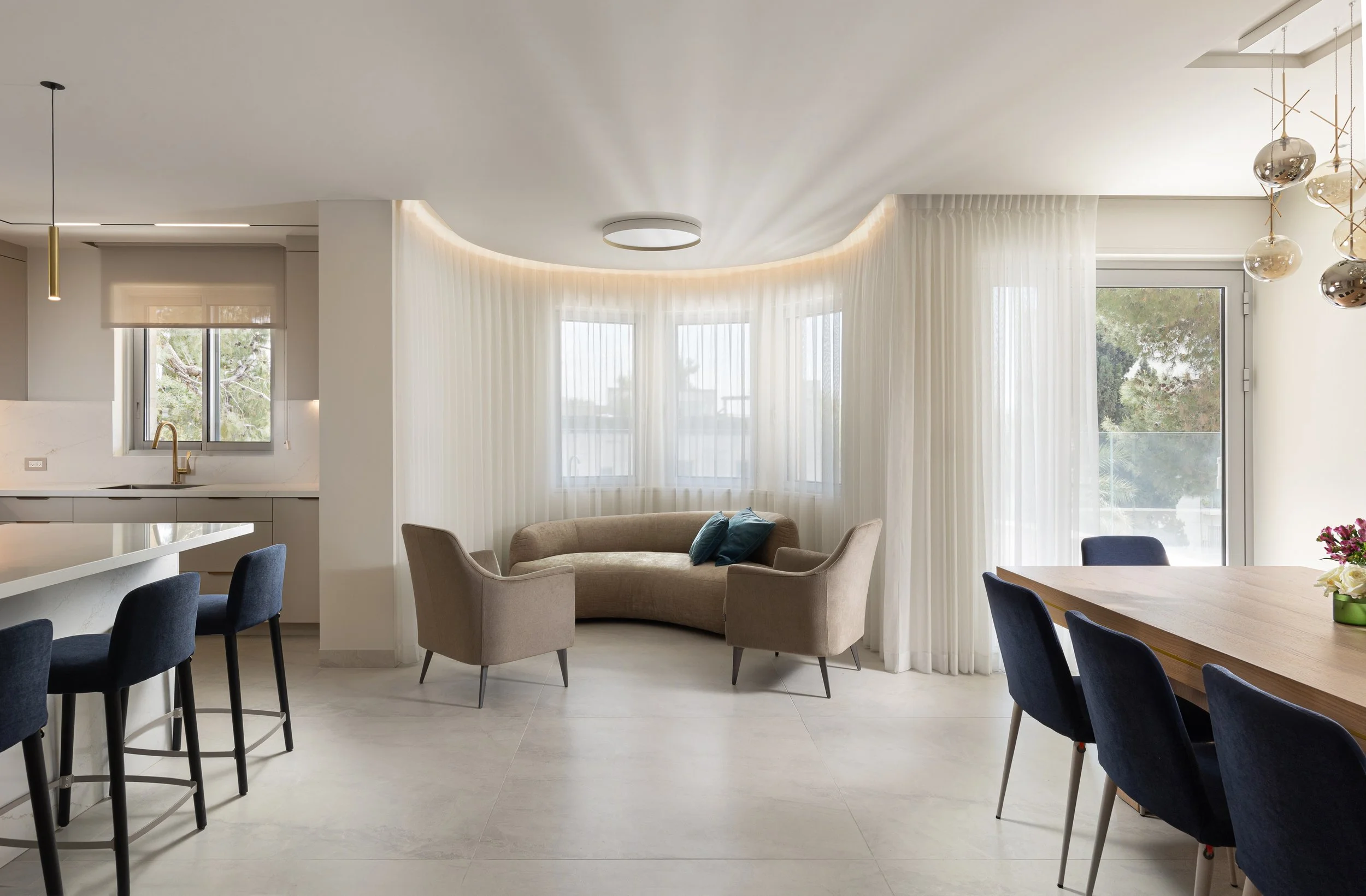 Modern interior of a living room and dining area with large windows, sheer curtains, a beige curved sofa with cushions, a round dining table with blue chairs, pendant lights, and a ceiling fixture, with trees visible outside.