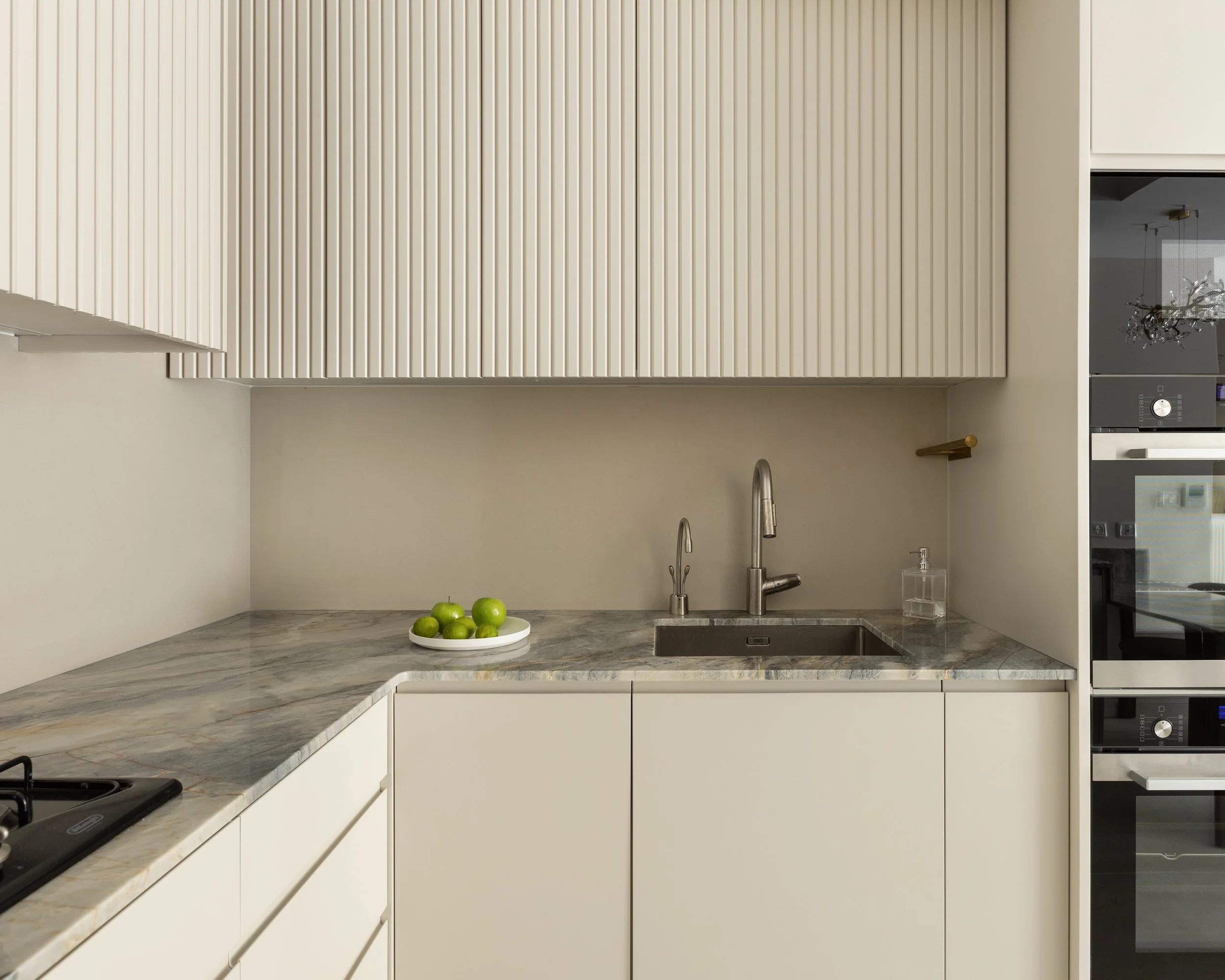 Modern kitchen with white cabinets, marble countertop, and a stainless steel sink; a bowl of green apples on the counter; beige walls; there's a window on the right side.