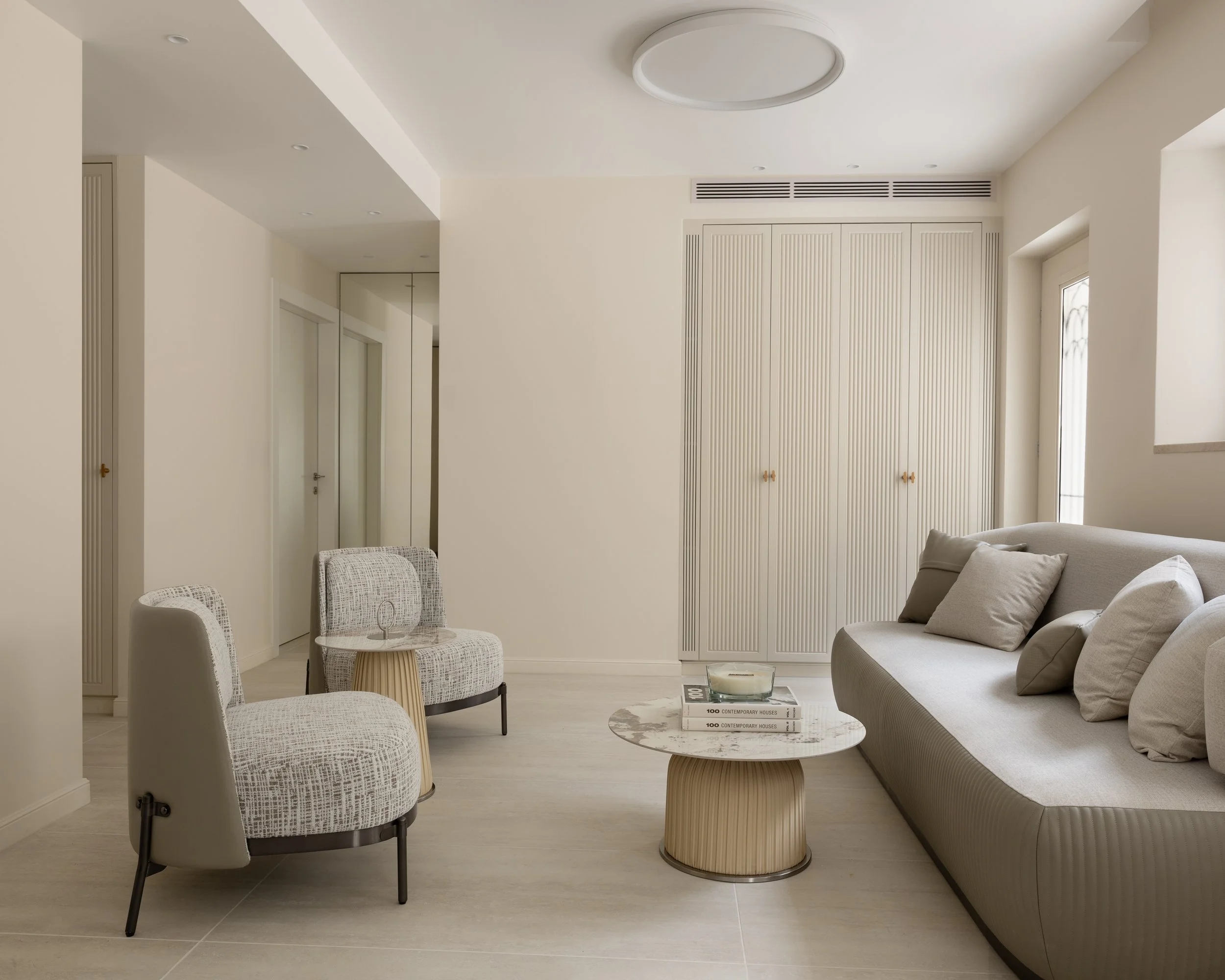 A modern, minimalistic living room with beige walls, a curved sofa, and a round coffee table. There are two patterned chairs and a stack of books on the coffee table. Natural light enters through a nearby window.
