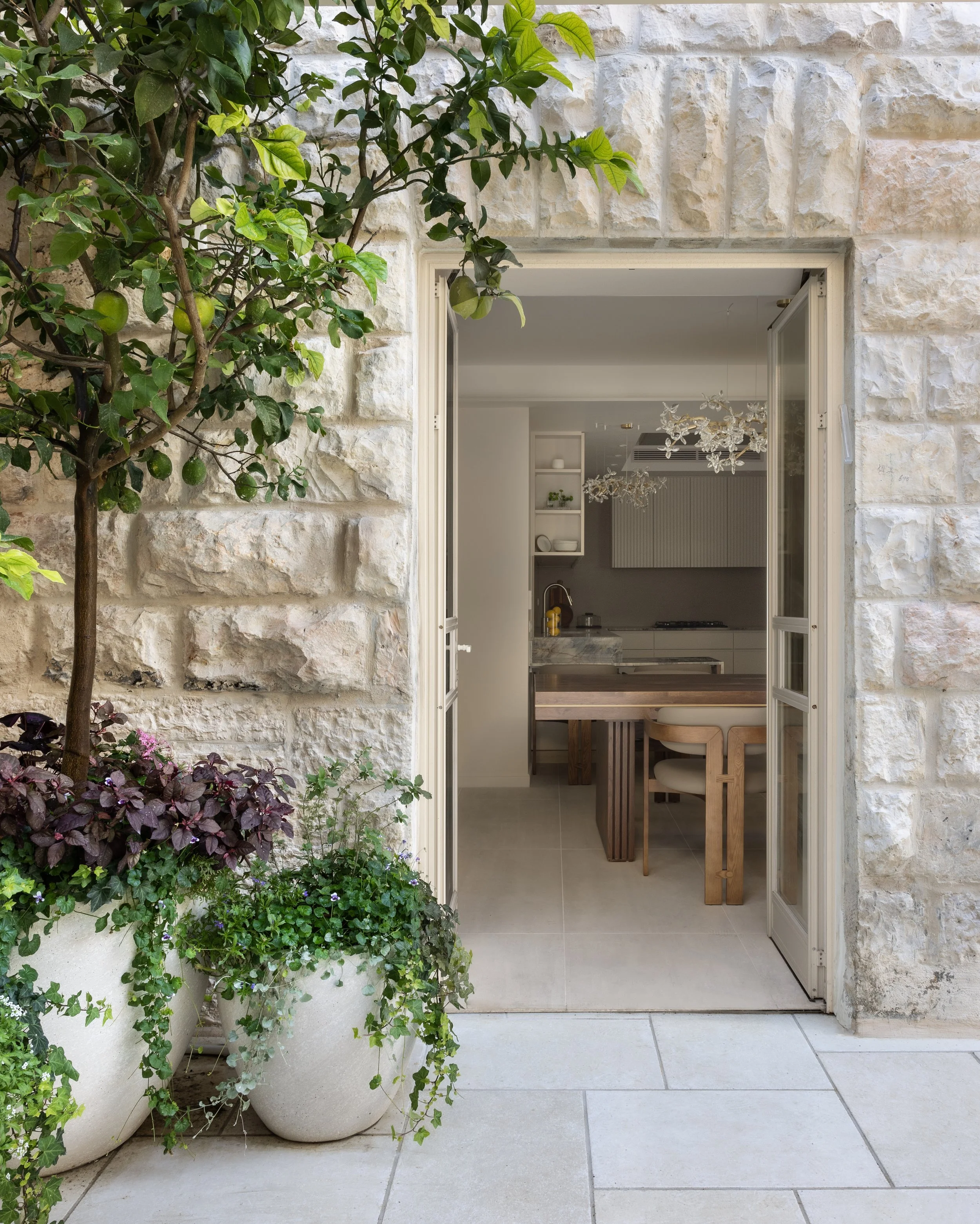 View of an open door leading from an outdoor patio with large potted plants on the left, into a bright interior room with a wooden table, chairs, shelves, and a modern chandelier.