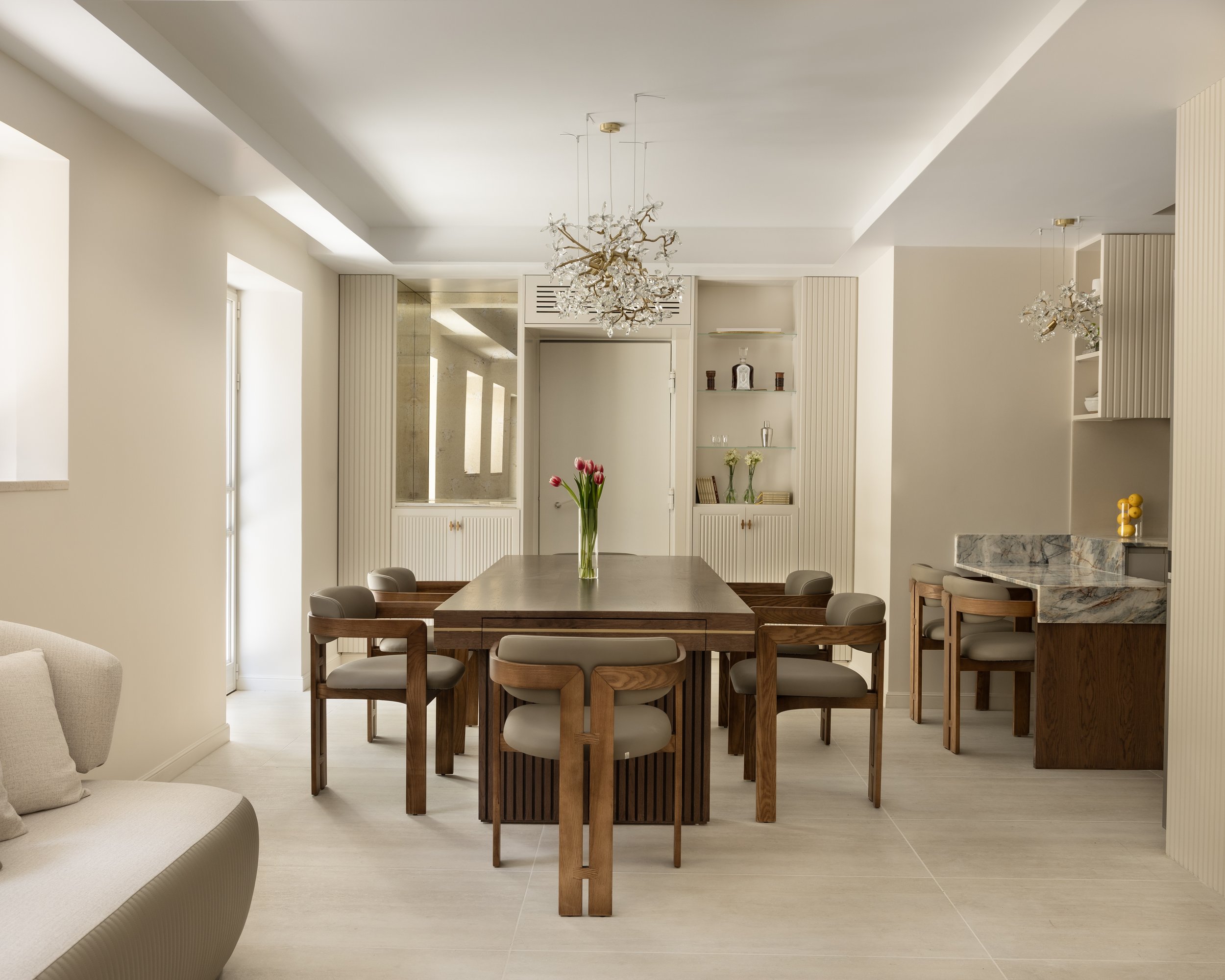 Modern dining room with a wooden dining table, beige chairs, and decorative chandeliers hanging from the ceiling. Bright natural light streams in through the windows.