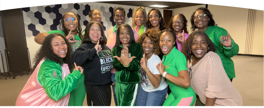 Group of women smiling and posing together indoors, some making hand gestures, with a hexagonal pattern wall in the background.