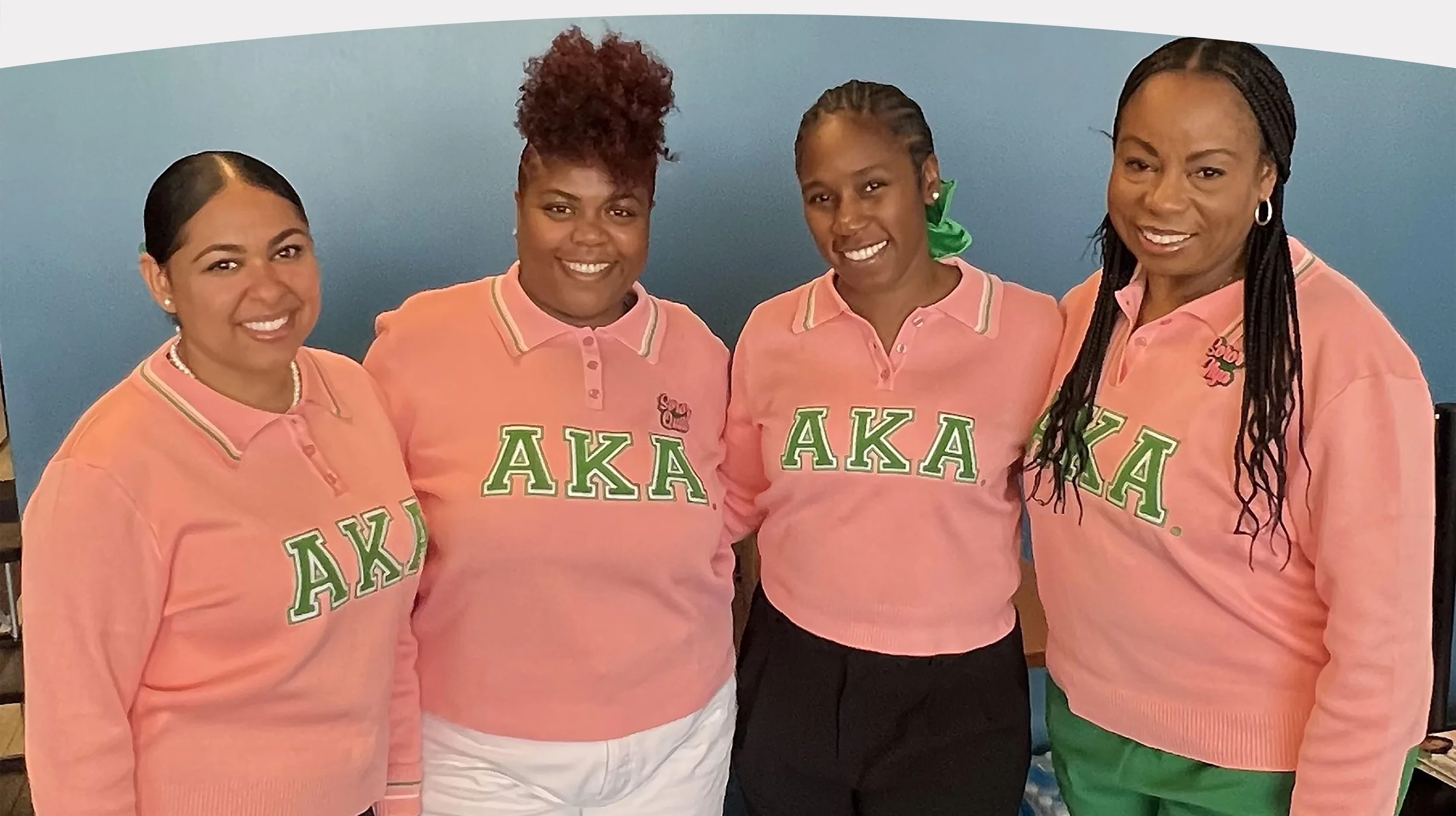 Four women wearing pink shirts with green Greek letters 'ΑΚΑ' on the front, smiling, standing together in front of a blue wall.