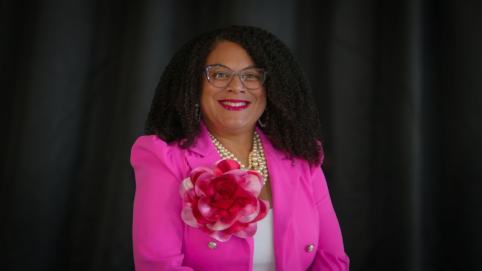 A woman with curly black hair, glasses, and bright pink lipstick, smiling at the camera, dressed in a pink blazer with a large pink flower accessory on the lapel, wearing pearl jewelry, against a dark background.