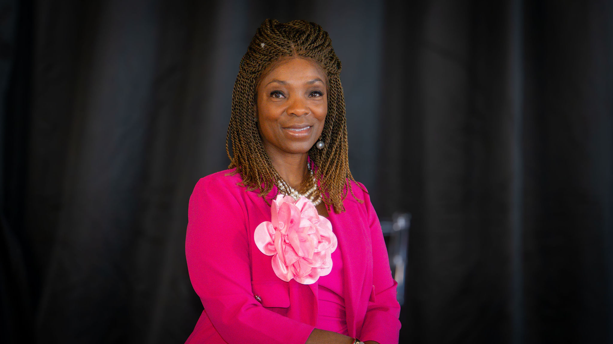 A woman with braided hair wearing a bright pink blazer and a large pink floral accessory on her chest, standing against a dark, blurred background.