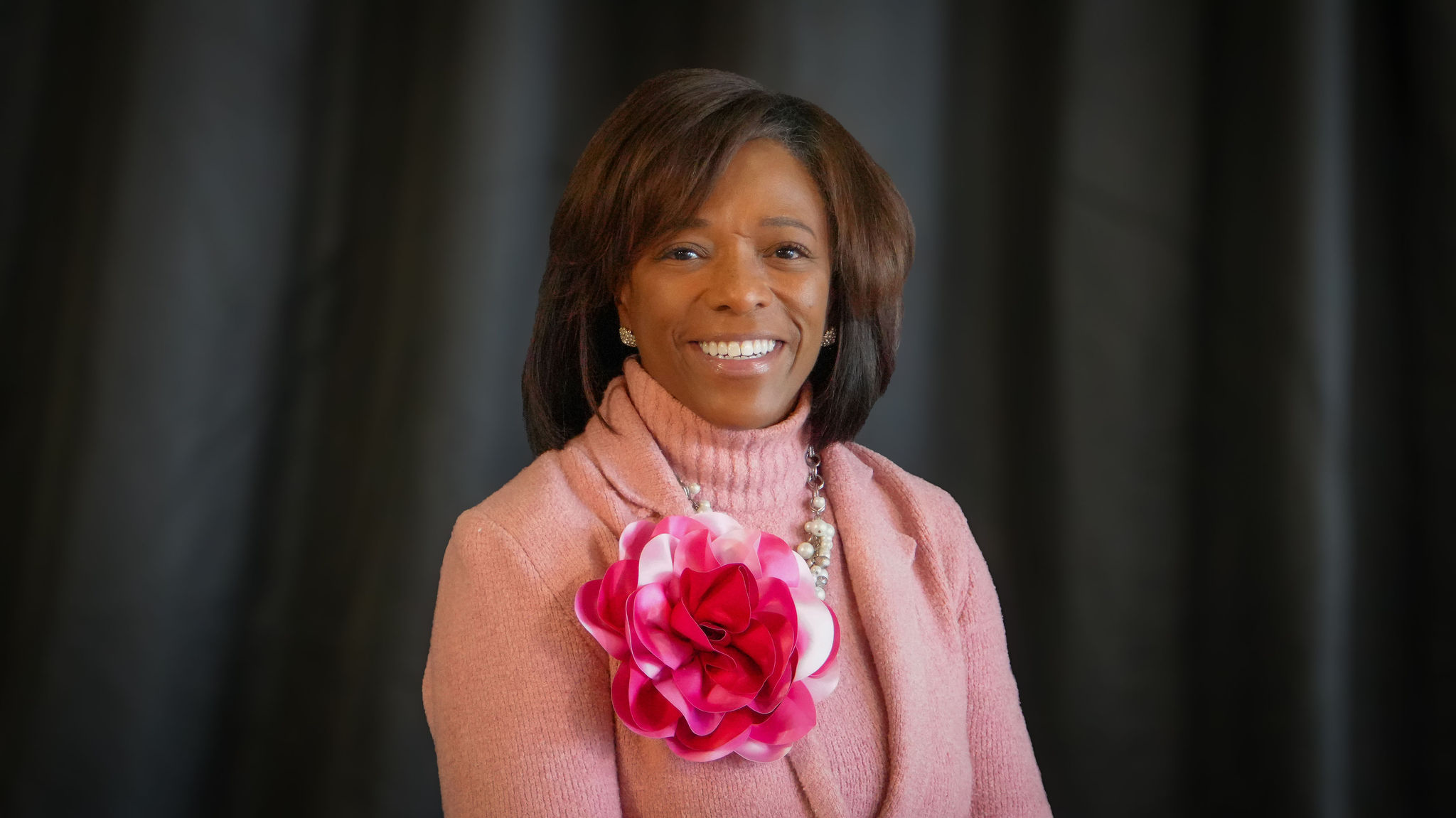 A smiling woman with shoulder-length brown hair wearing a pink sweater and a large pink and white flower brooch, standing against a dark background.