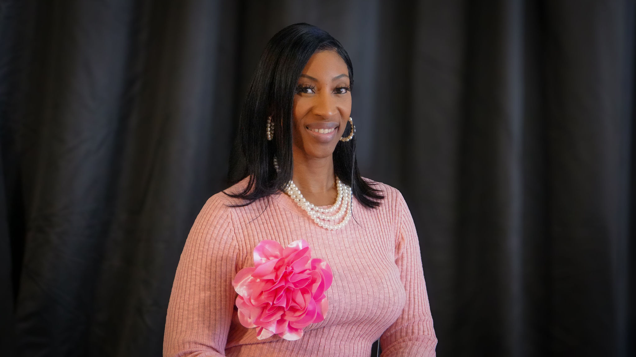 A woman with long black hair, wearing a pink ribbed sweater, pearl earrings, and a pearl necklace, stands in front of a dark curtain. She is smiling and holding a large pink fabric flower.