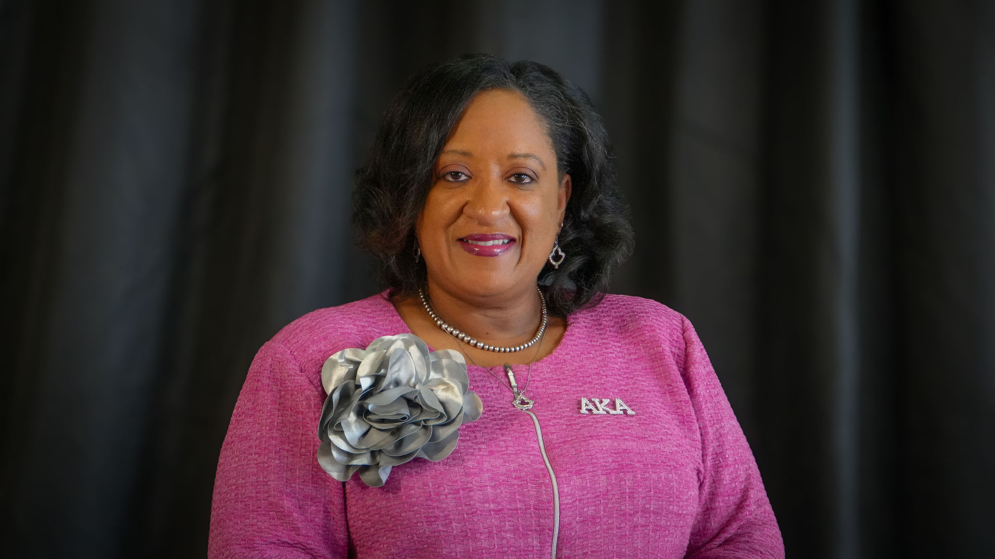 A woman with dark wavy hair, wearing a pink dress with a silver and gray flower accessory on her left shoulder. She has a necklace and earrings, and is smiling against a dark background.
