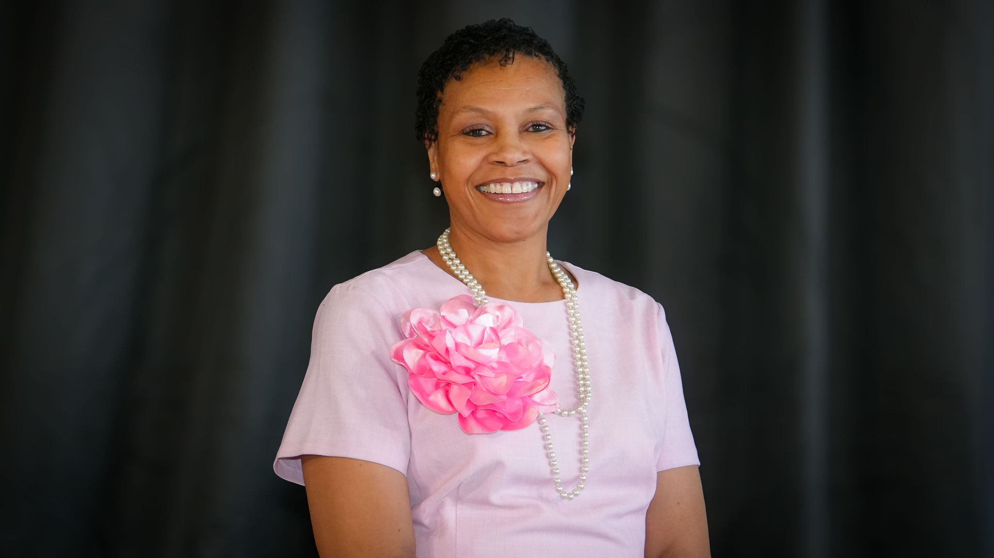 A woman with short curly hair smiling, wearing a pink dress with a large pink fabric flower and pearl necklace, against a dark background.