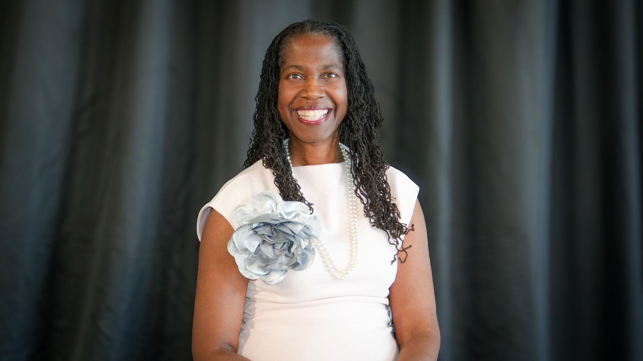A smiling African American woman with long curly hair, dressed in a white dress with a silver flower detail on the shoulder, wearing a pearl necklace, standing in front of dark curtains.