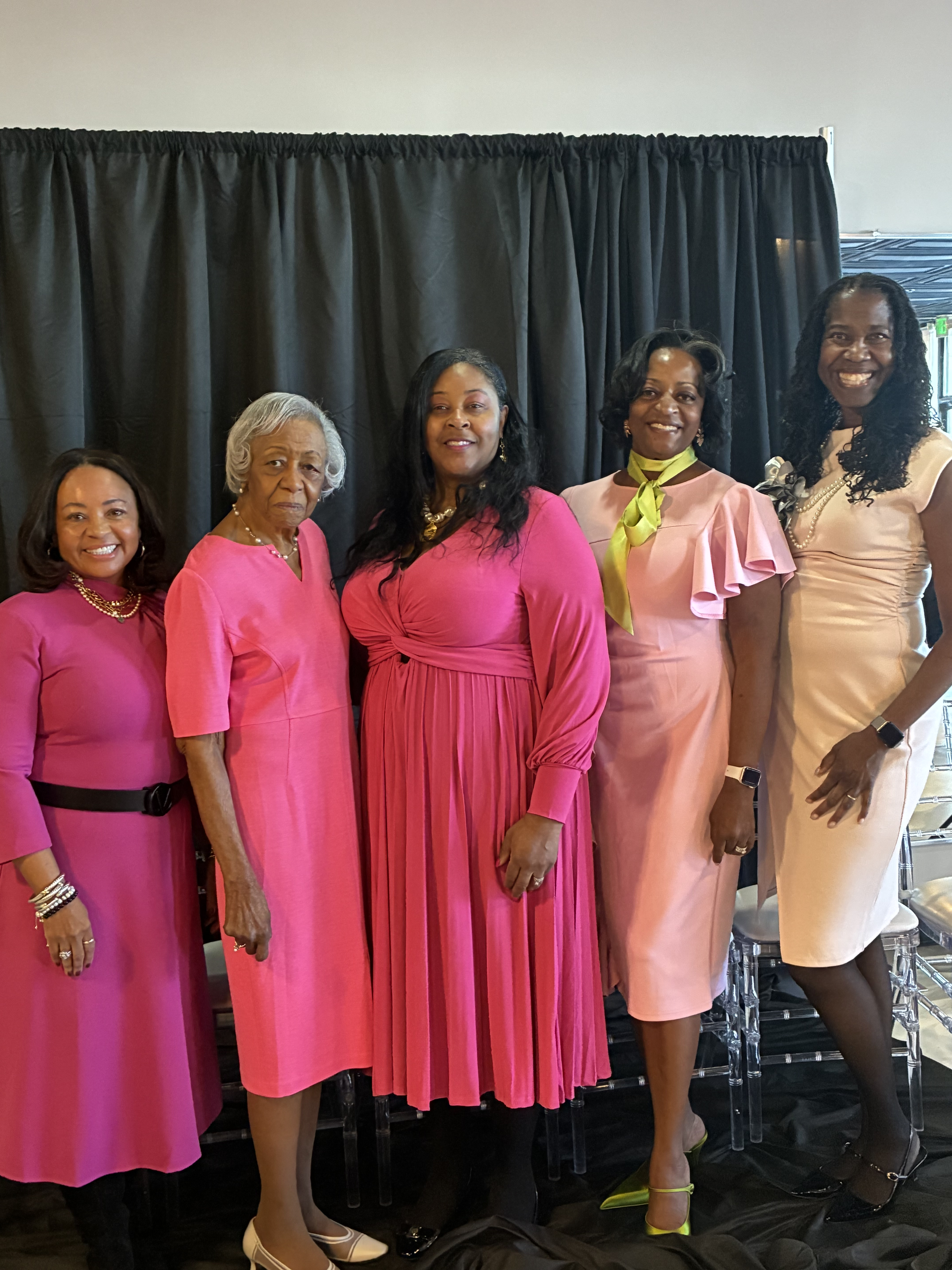 Five women dressed in pink dresses standing in front of a black curtain at an indoor event.