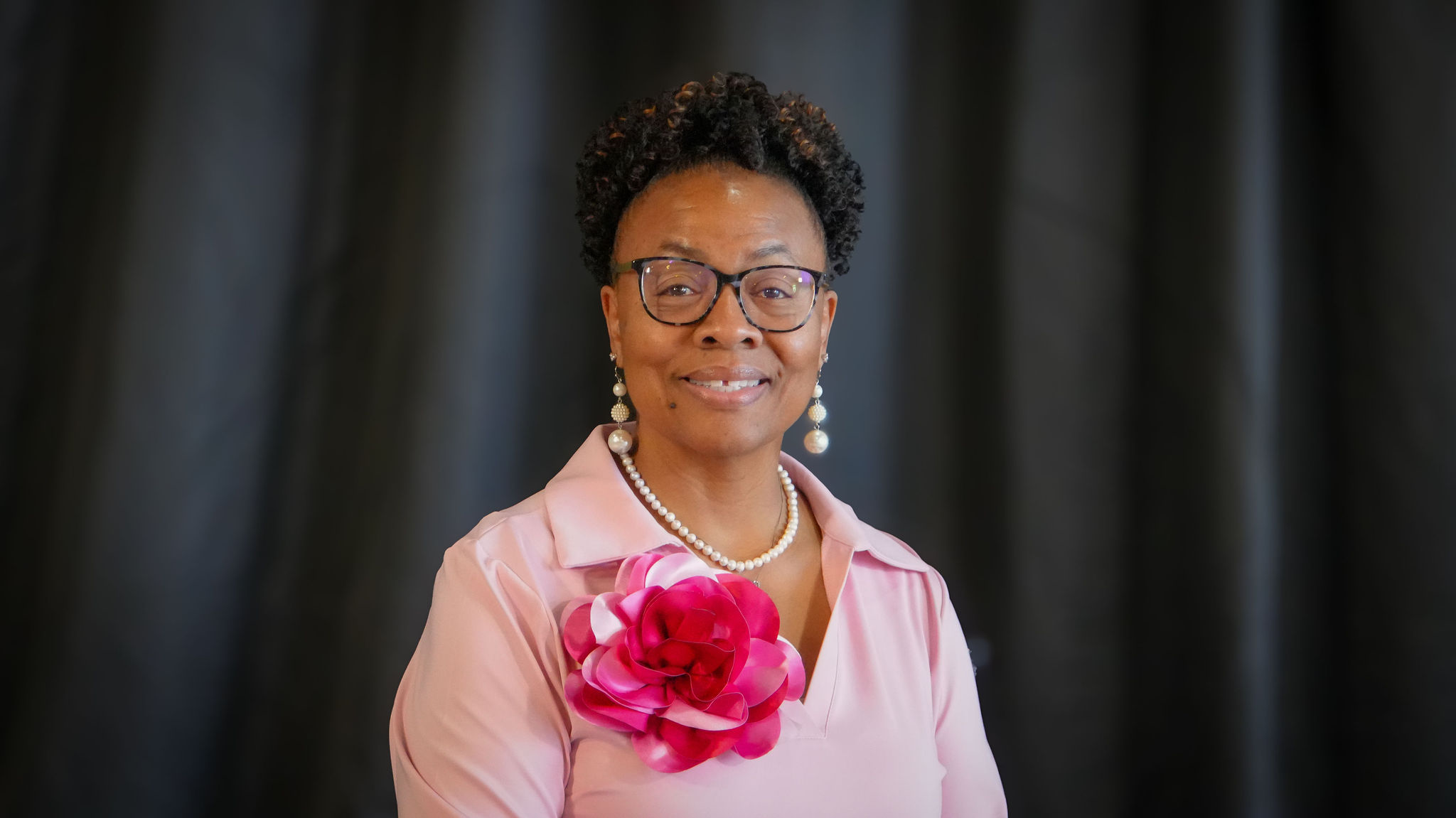 A woman with short curly black hair, glasses, pearl jewelry, and a pink outfit with pink and red flower decorations on her chest, standing in front of a dark background.