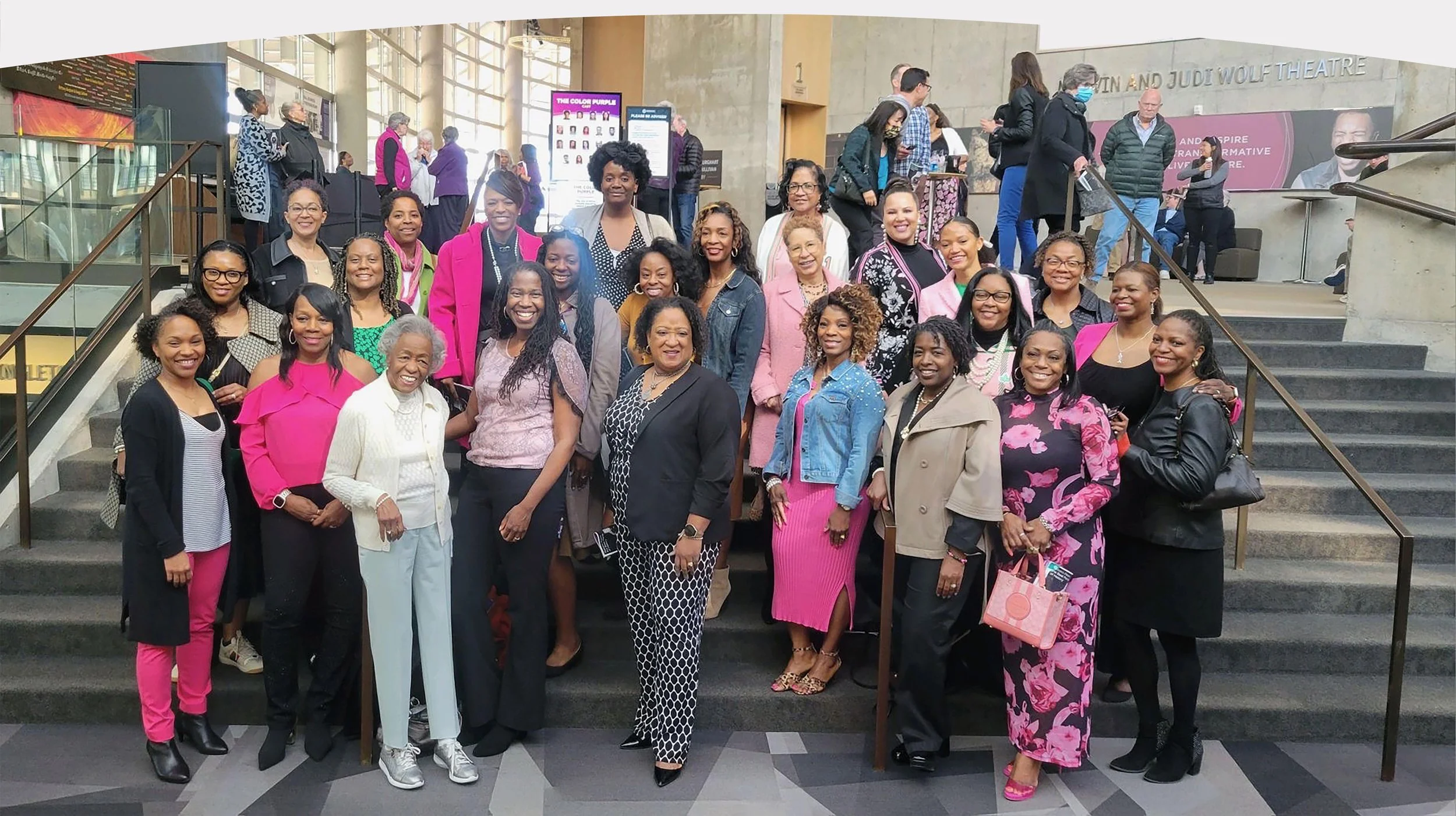 A diverse group of women gathered on the steps of a theater, smiling for a photo. The background shows other people mingling and signs in a large indoor space.