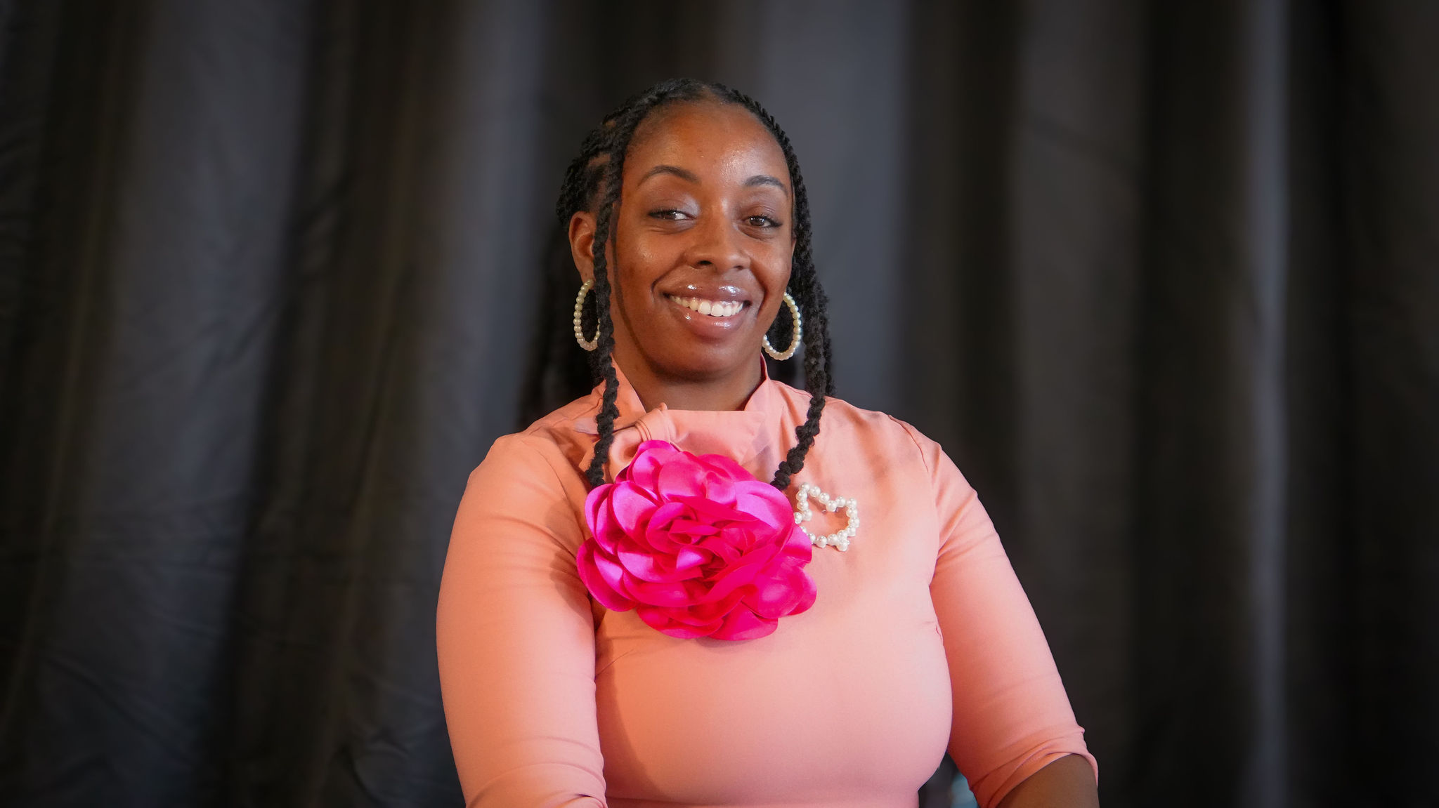 A smiling Black woman with braids wearing pearl earrings, a pink dress with a large pink fabric flower and a pearl necklace, standing in front of a dark, textured background.